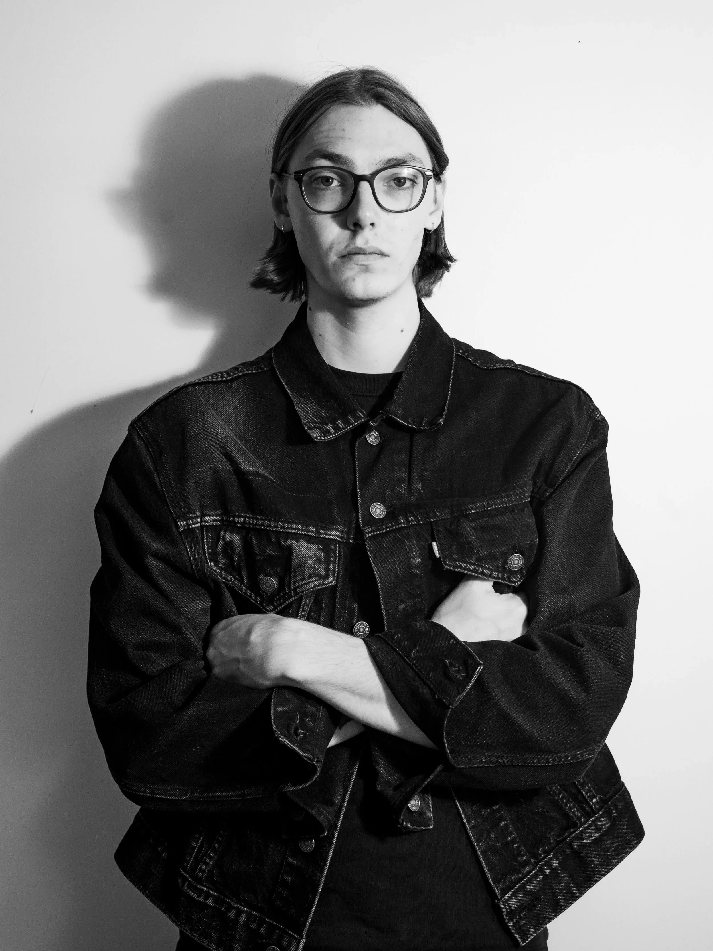 Black and white portrait of a person with shoulder-length hair, wearing glasses and a denim jacket, standing against a plain wall.
