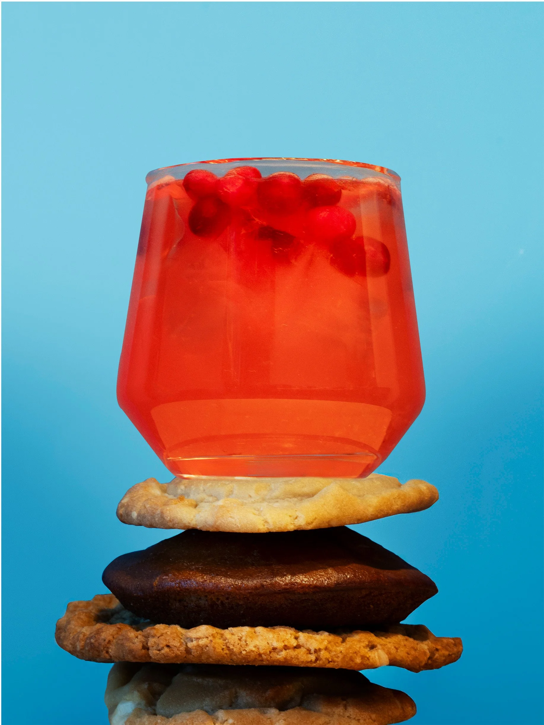 A glass of pink beverage with berries on top, balanced on a stack of rocks and cookies against a blue background.