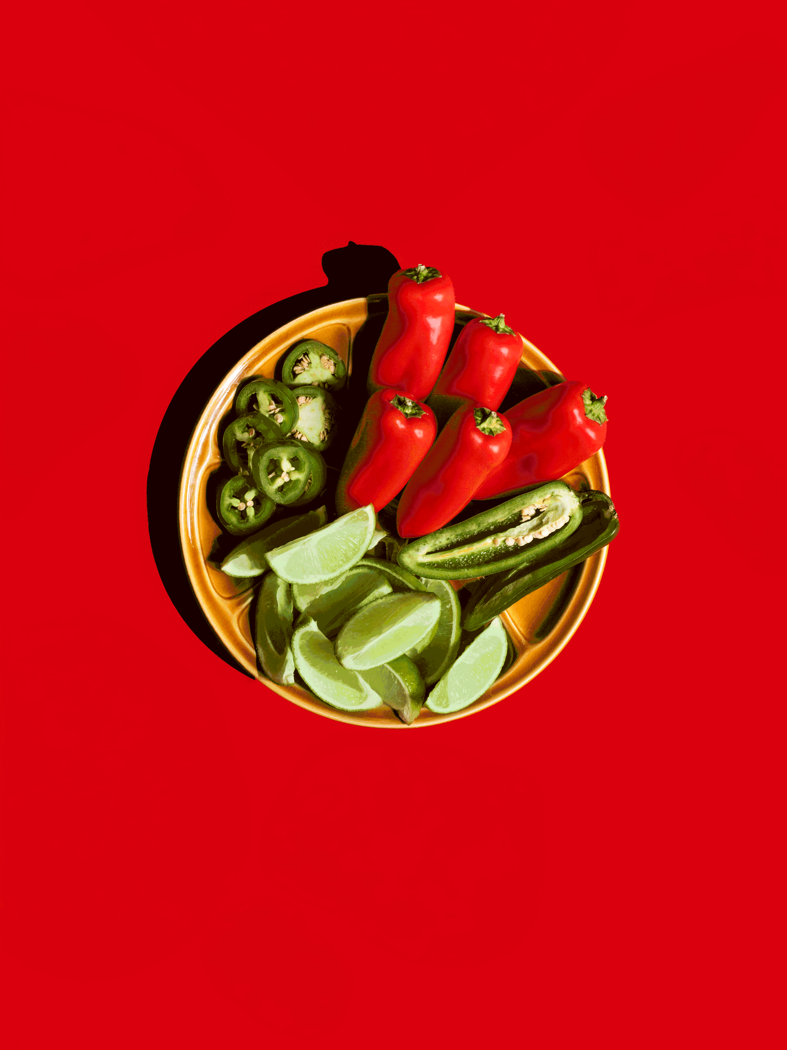 A top view of a round yellow bowl filled with sliced green jalapeño peppers, lime wedges, and red jalapeño peppers on a red background.