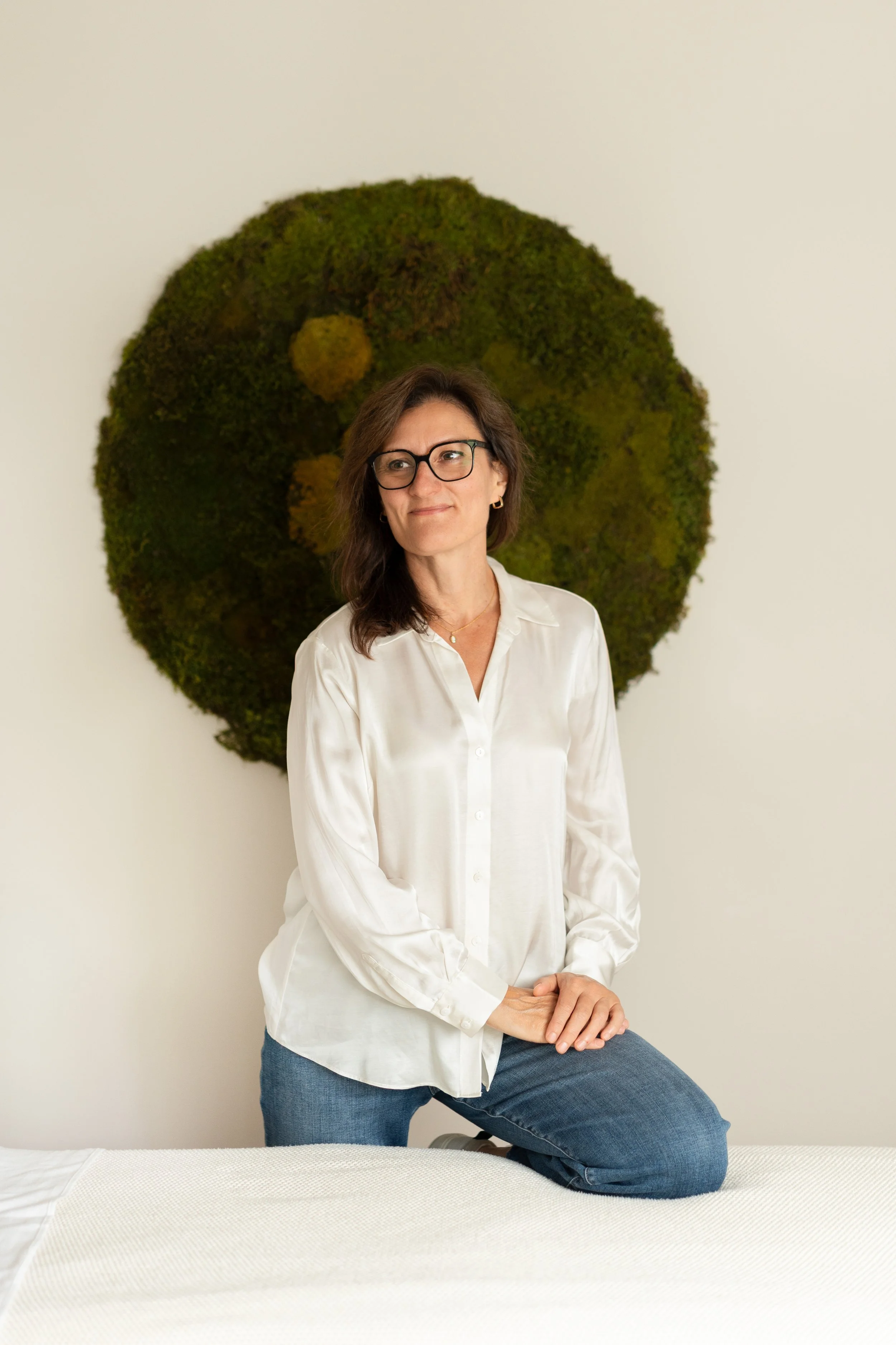 A woman with glasses, wearing a white shirt and jeans, sitting on a bed in front of a large moss art piece on a white wall.