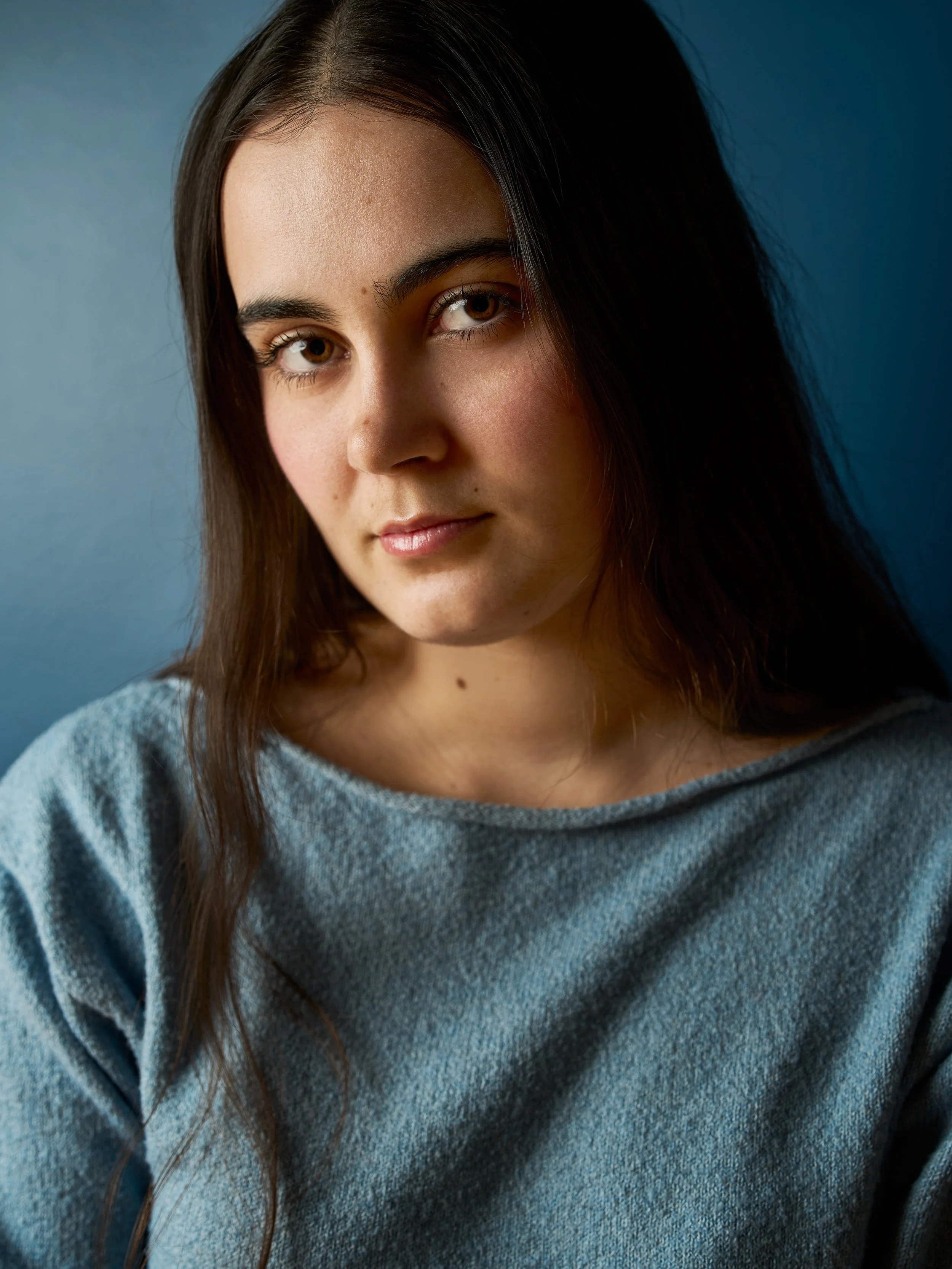 Close-up portrait of a young woman with long dark hair, wearing a gray sweatshirt, against a blue background.