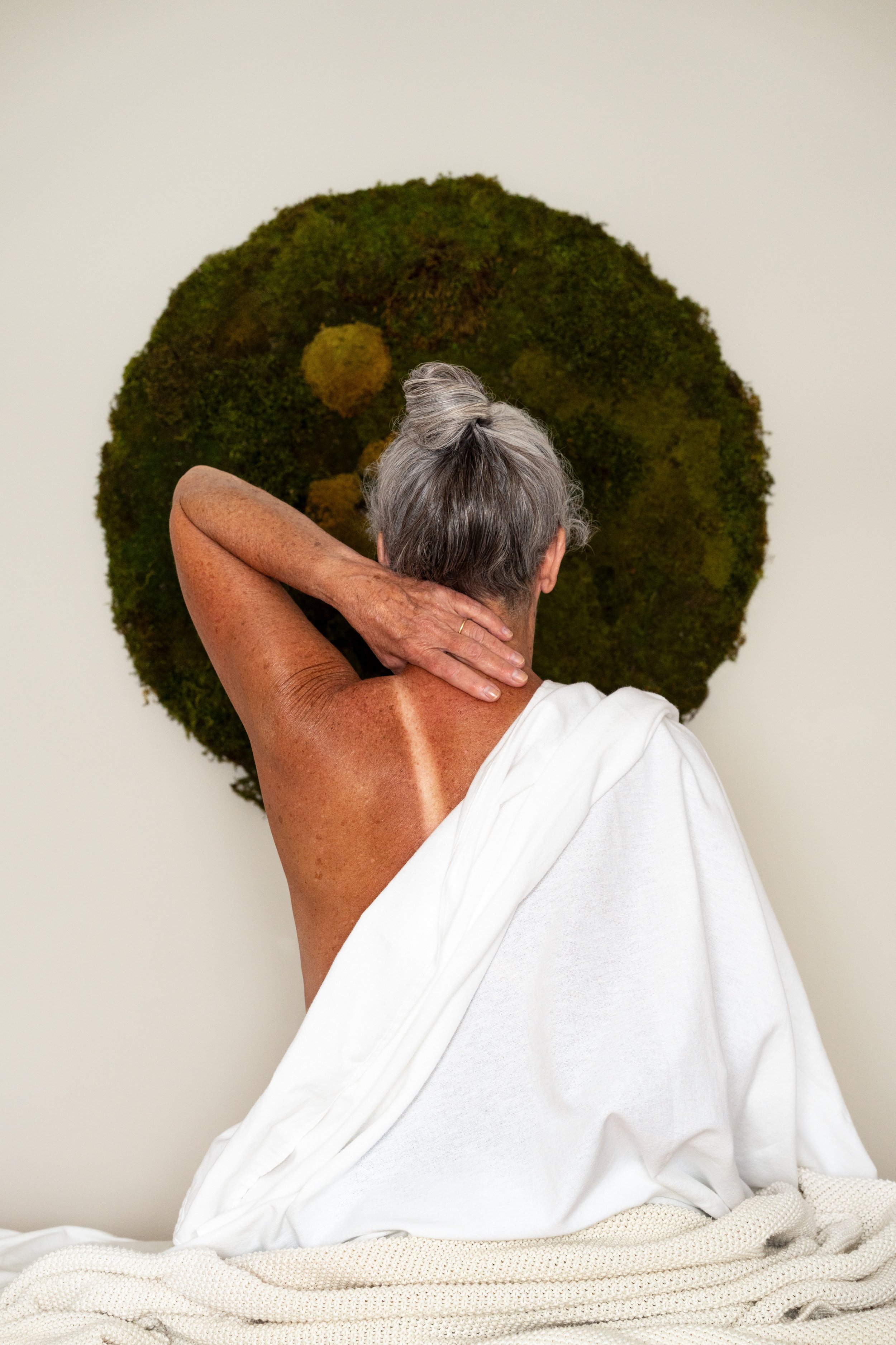 A person with gray hair, seen from behind, resting their head on one arm while seated. They are partially covered with a white cloth and are in front of a textured, moss-covered circular artwork or sculpture on a plain wall.