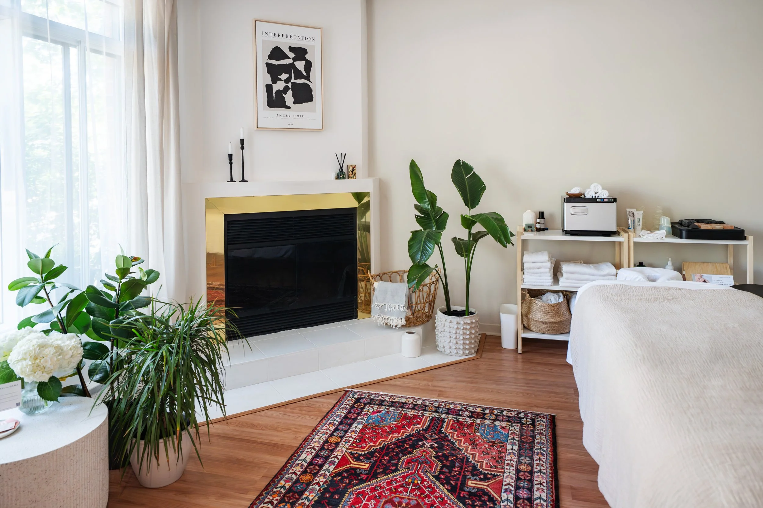 A cozy living space with white walls, a fireplace with a black and gold frame, and a large window with sheer white curtains. There are green plants, a colorful oriental rug on wooden flooring, and a white bed partially visible on the right. A shelf holds rolled towels, skincare products, and a sterilizer, with additional plants and decor around.