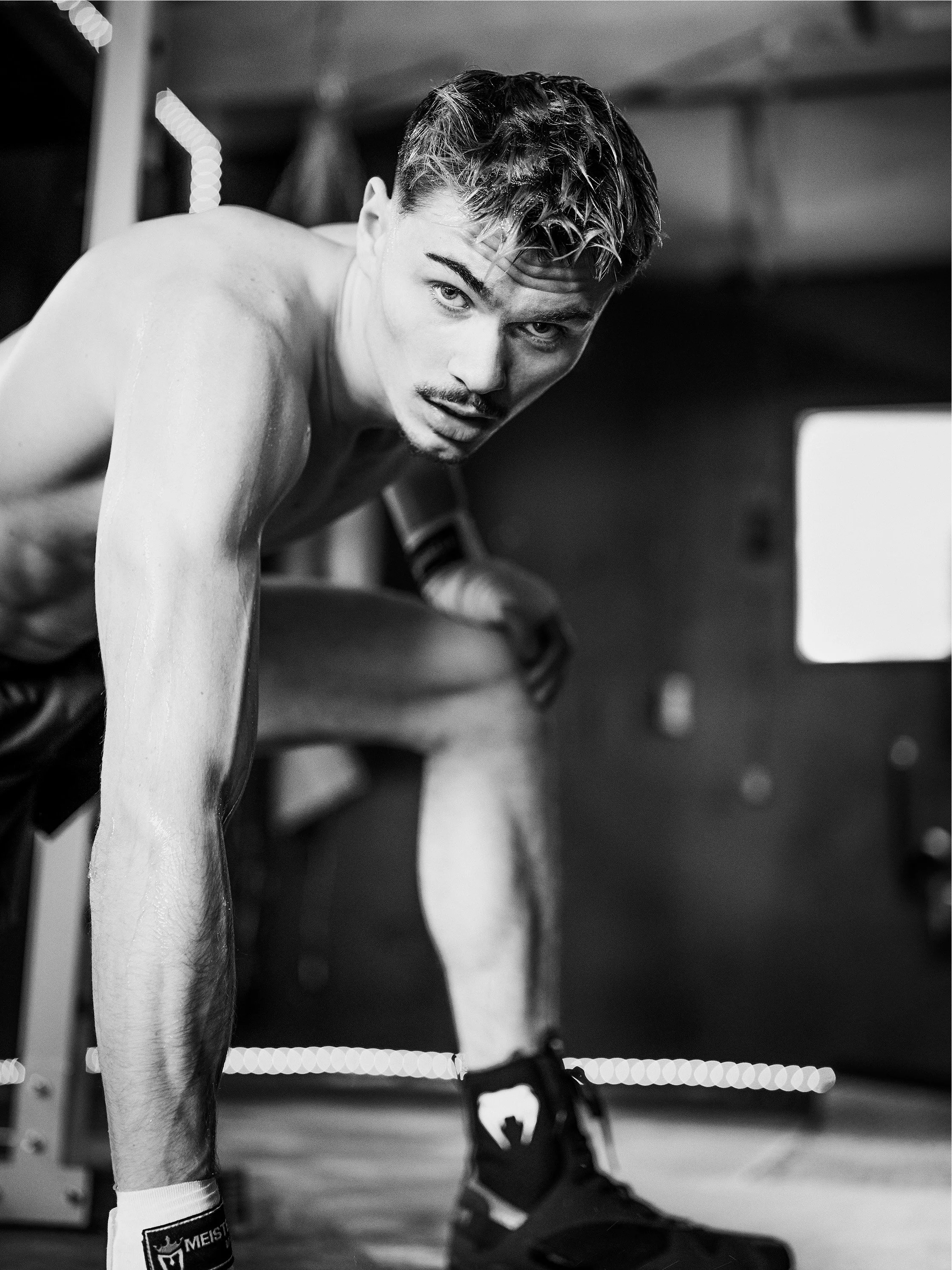 Black and white photo of a shirtless man with wet hair in a gym, leaning forward with one hand on the floor and the other resting on his knee, looking towards the camera.