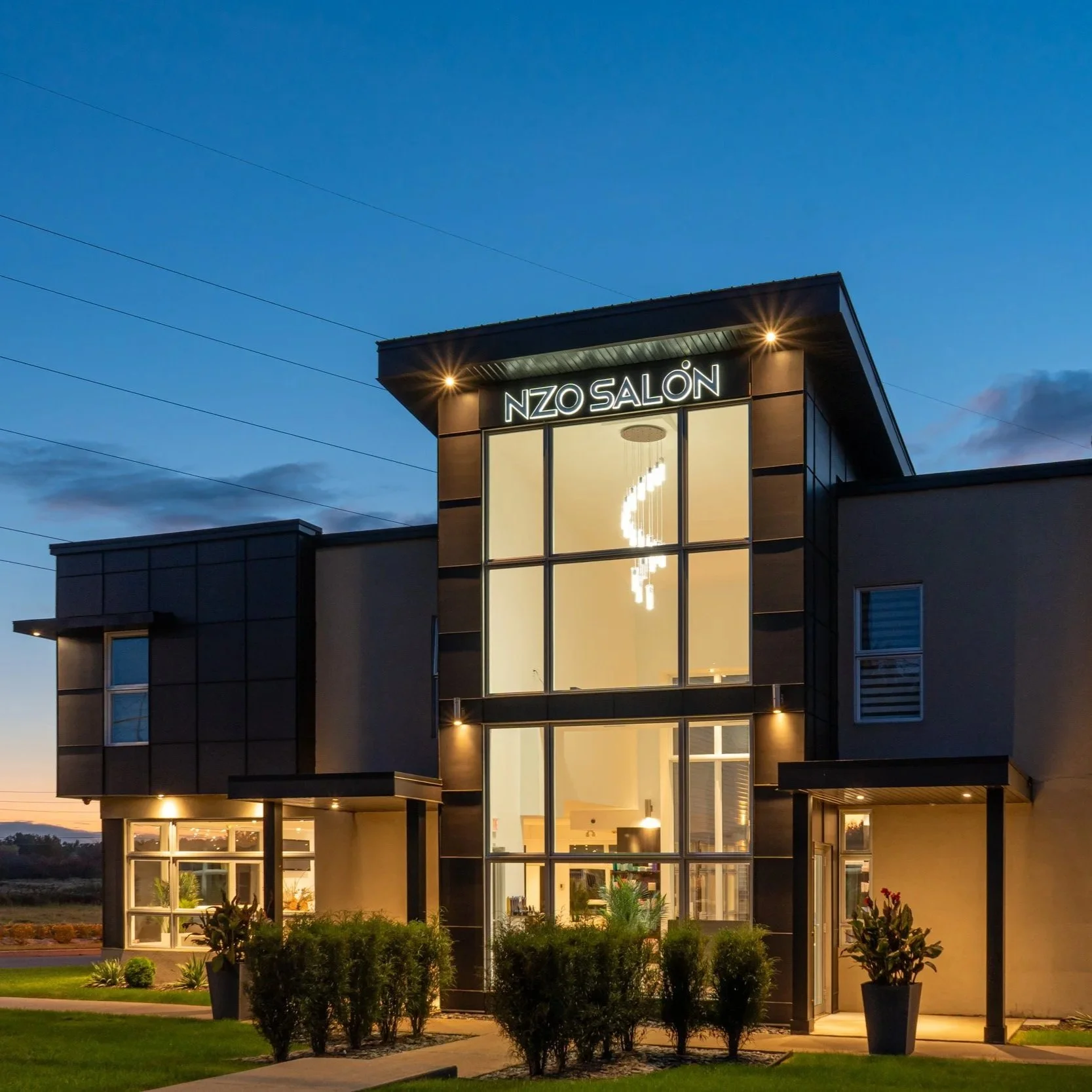 Modern commercial building with large glass windows and neon sign that reads 'NZO SALÓN' at dusk. Exterior lighting highlights the structure, and there are landscaped bushes and a lawn in front.