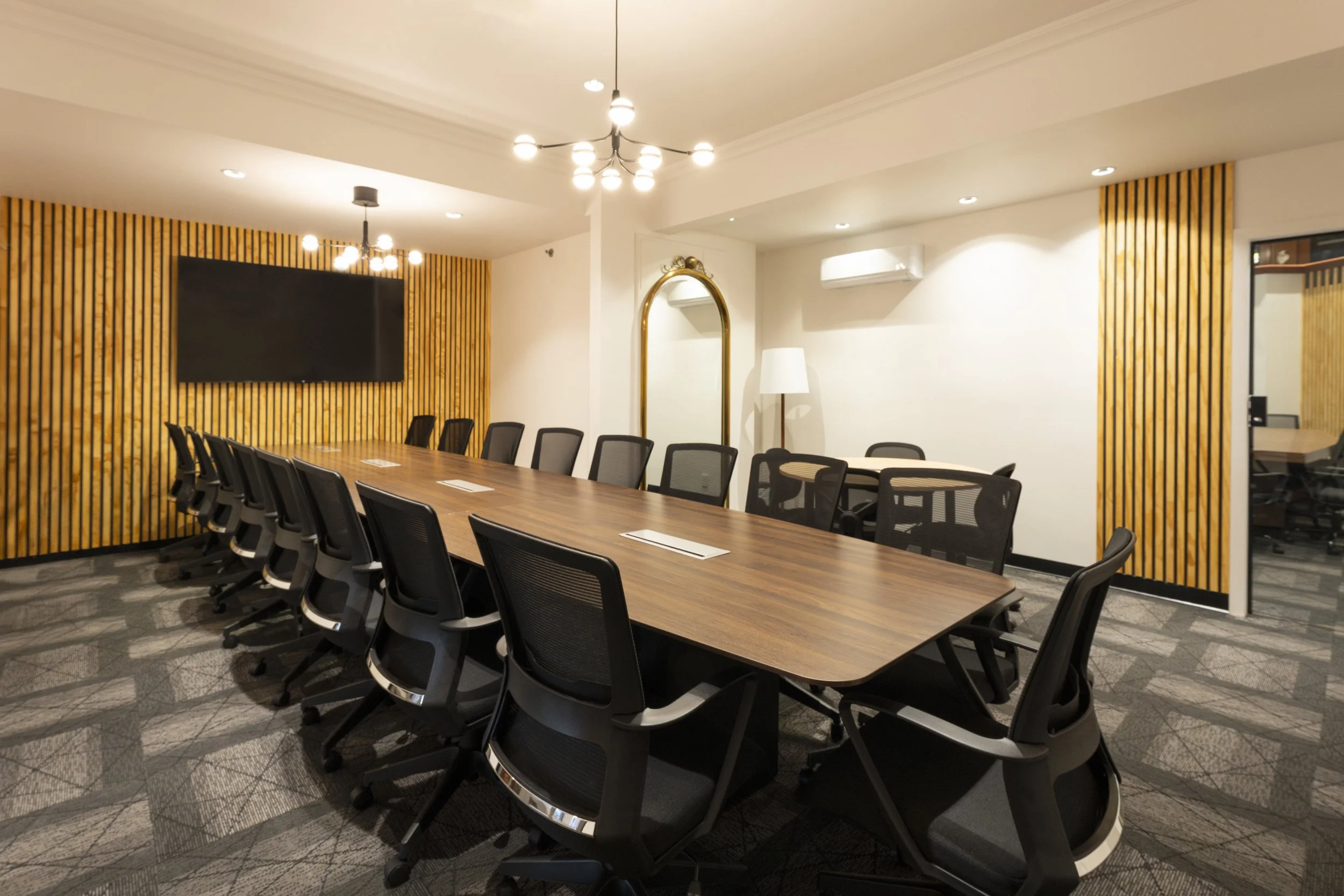 A modern conference room with a long wooden table surrounded by black office chairs, a large wall-mounted flat screen TV, wood panel accents, a white air conditioning unit, a standing white lamp, and minimalistic ceiling lighting fixtures.