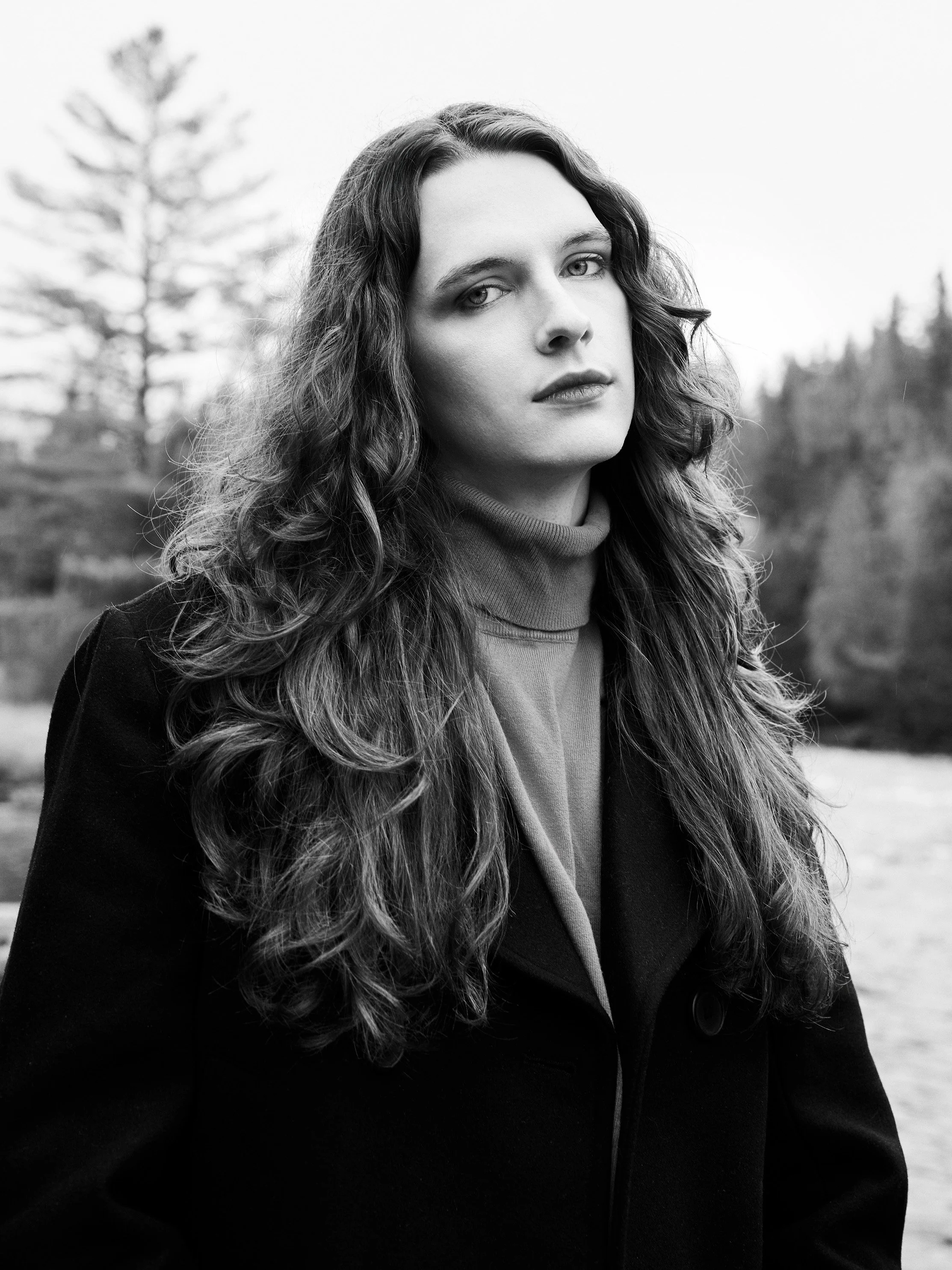 Black and white photo of a woman with long curly hair, wearing a jacket and a turtleneck, standing outdoors with trees in the background.