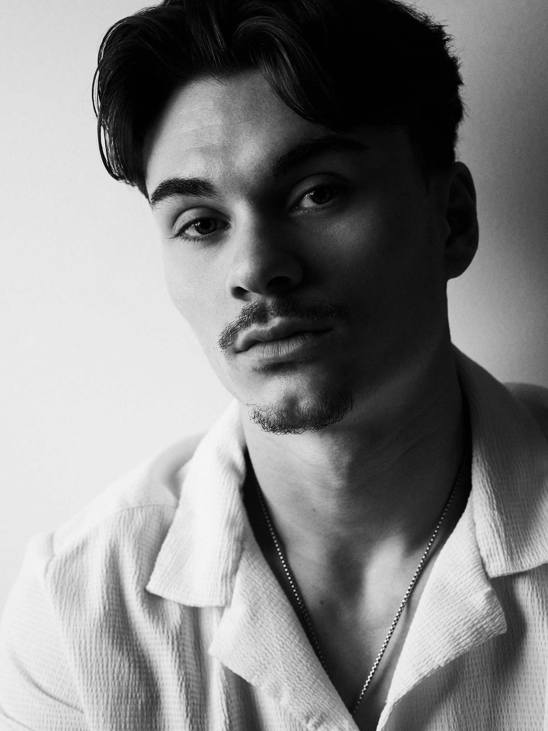 Black and white portrait of a young man with dark hair, light skin, wearing a white zip-up jacket and a chain necklace, looking into the camera.