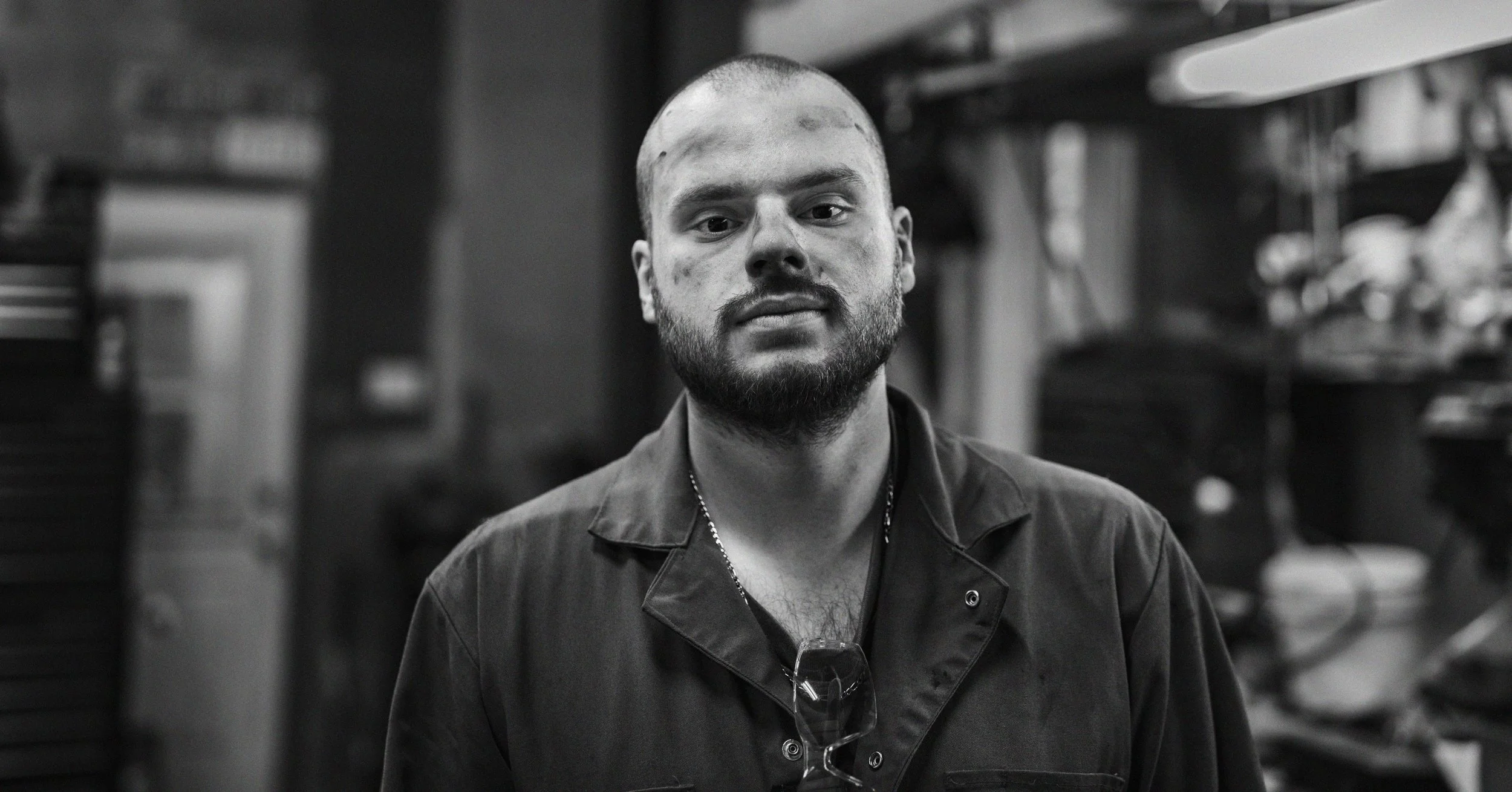 A young man with a beard and short hair, wearing a dark jacket and glasses hanging from a chain around his neck, stands in a room with shelves filled with various items. The image is in black and white.