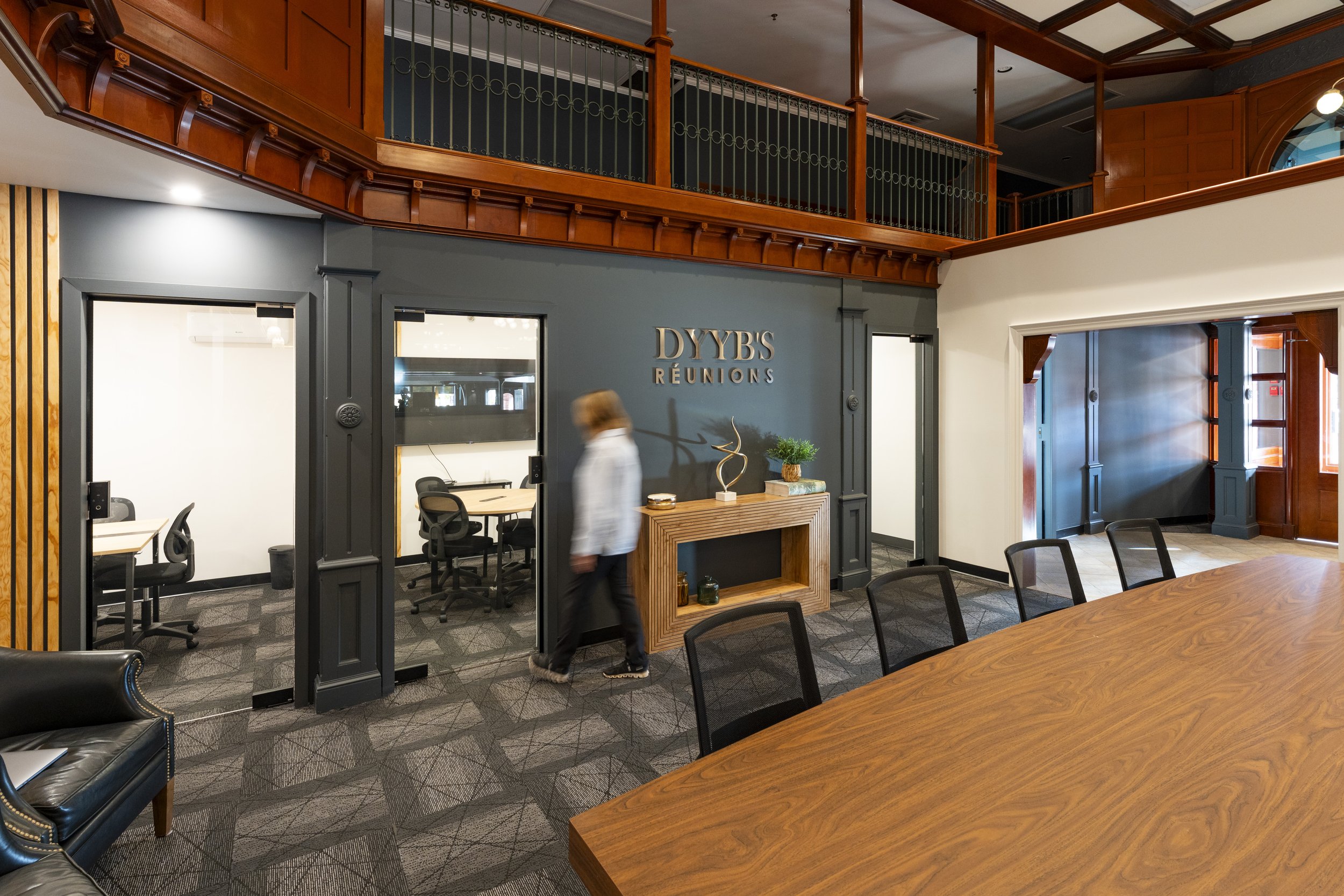 Interior of a modern office with wooden accents, a conference table, chairs, and a sign that reads 'DYYBS REUNIONS.' There is a person walking past a small decor shelf and a large wall-mounted TV.