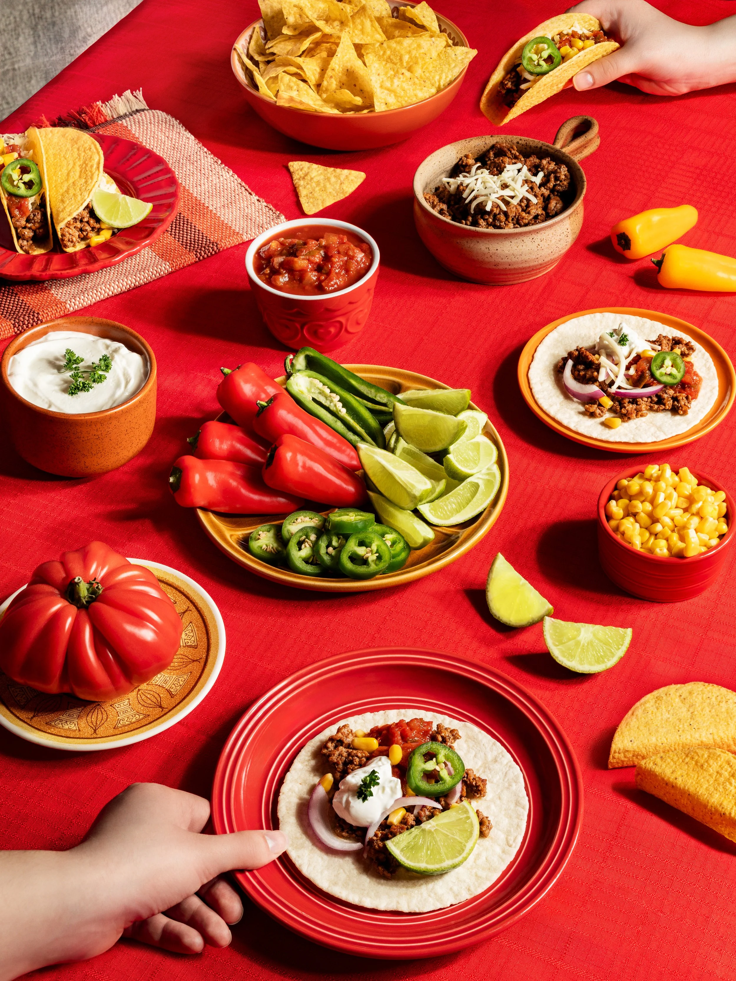 Person holding a taco with seasoned ground meat, sliced jalapeños, onions, corn, and a lime wedge on a red plate. Other tacos, bowls of ground beef with cheese, salsa, sour cream, tortilla chips, and fresh vegetables like tomatoes, jalapeños, and lime slices are on a red tablecloth.