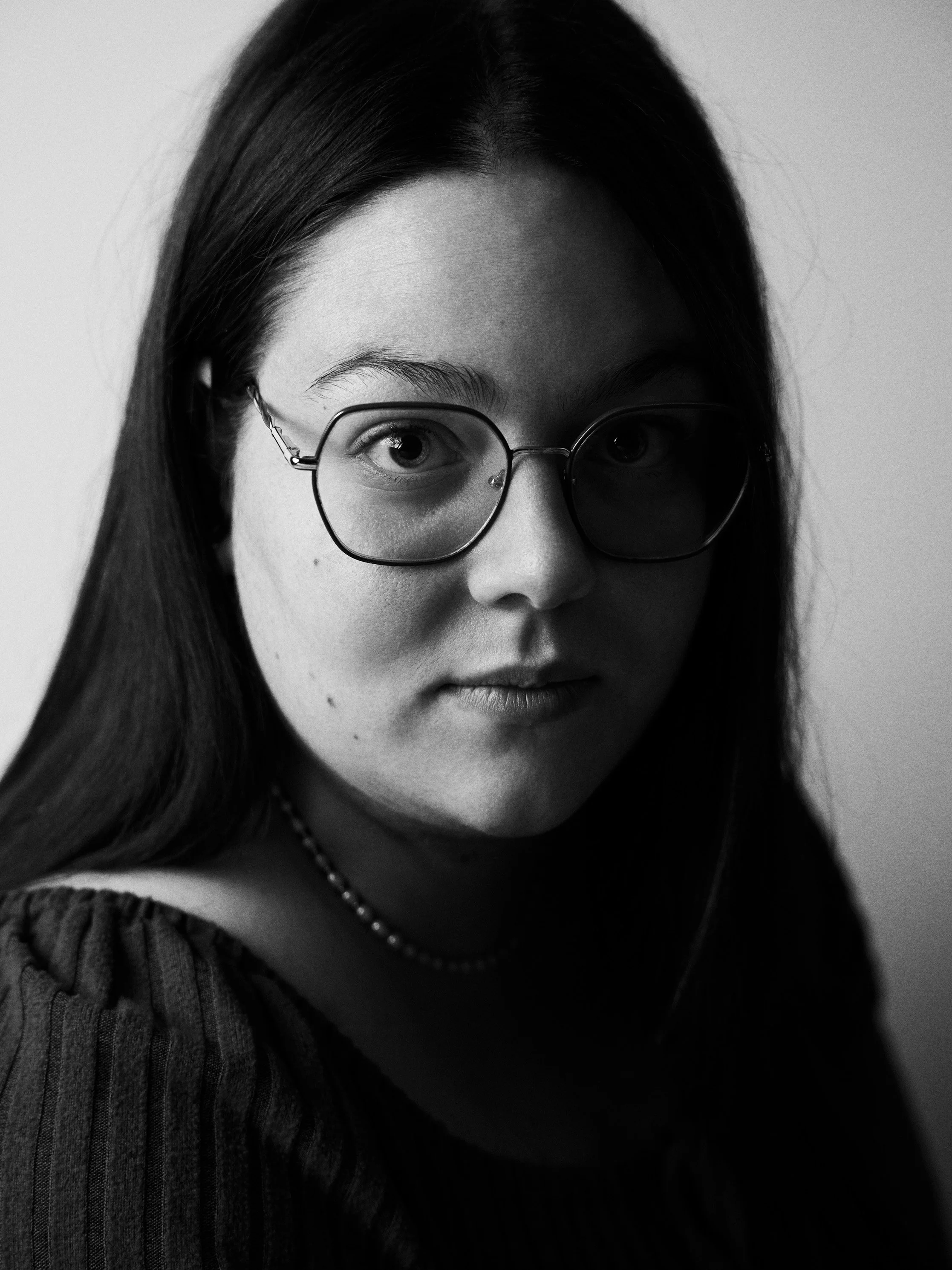 Close-up black and white portrait of a young woman with long dark hair wearing glasses, a necklace, and a sweater.