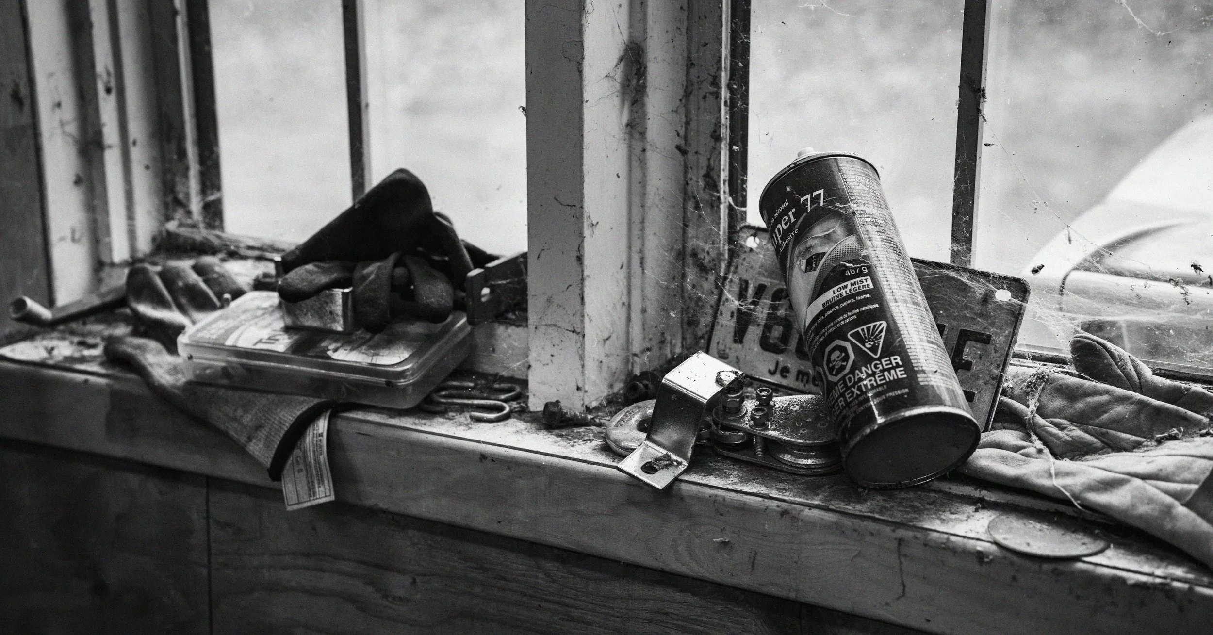 A cluttered window sill with dirty gloves, a spray can labeled 'Extreme' with safety warnings, a small device, a worn-out sign, and various small objects, all covered in dust and cobwebs.