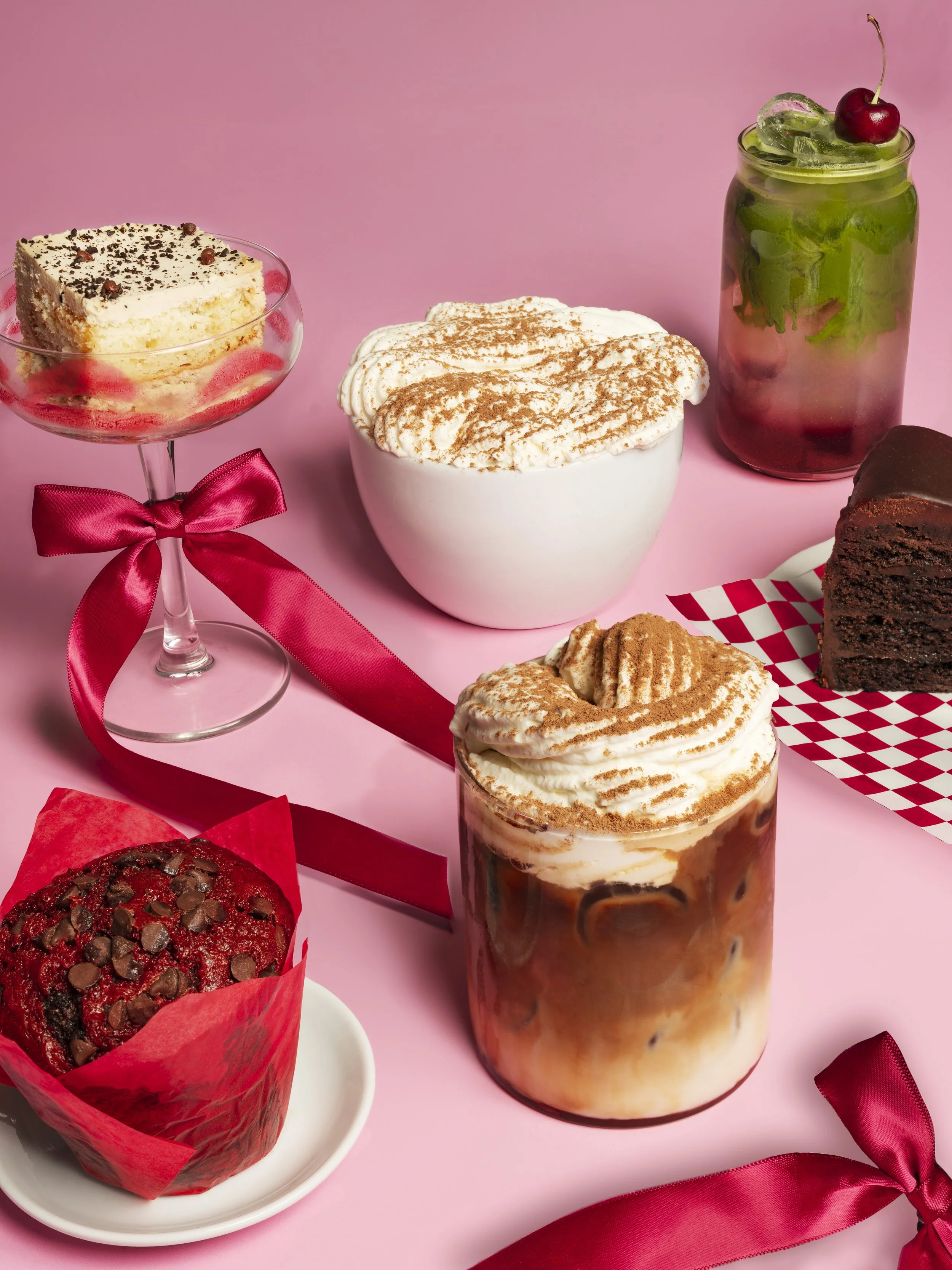 Various desserts and a beverage on a pink surface, including a slice of cake with chocolate shavings, a bowl of whipped cream with cinnamon, a jar of iced coffee with whipped cream, a chocolate cupcake with chocolate chips, and a slice of chocolate cake, decorated with red ribbons.