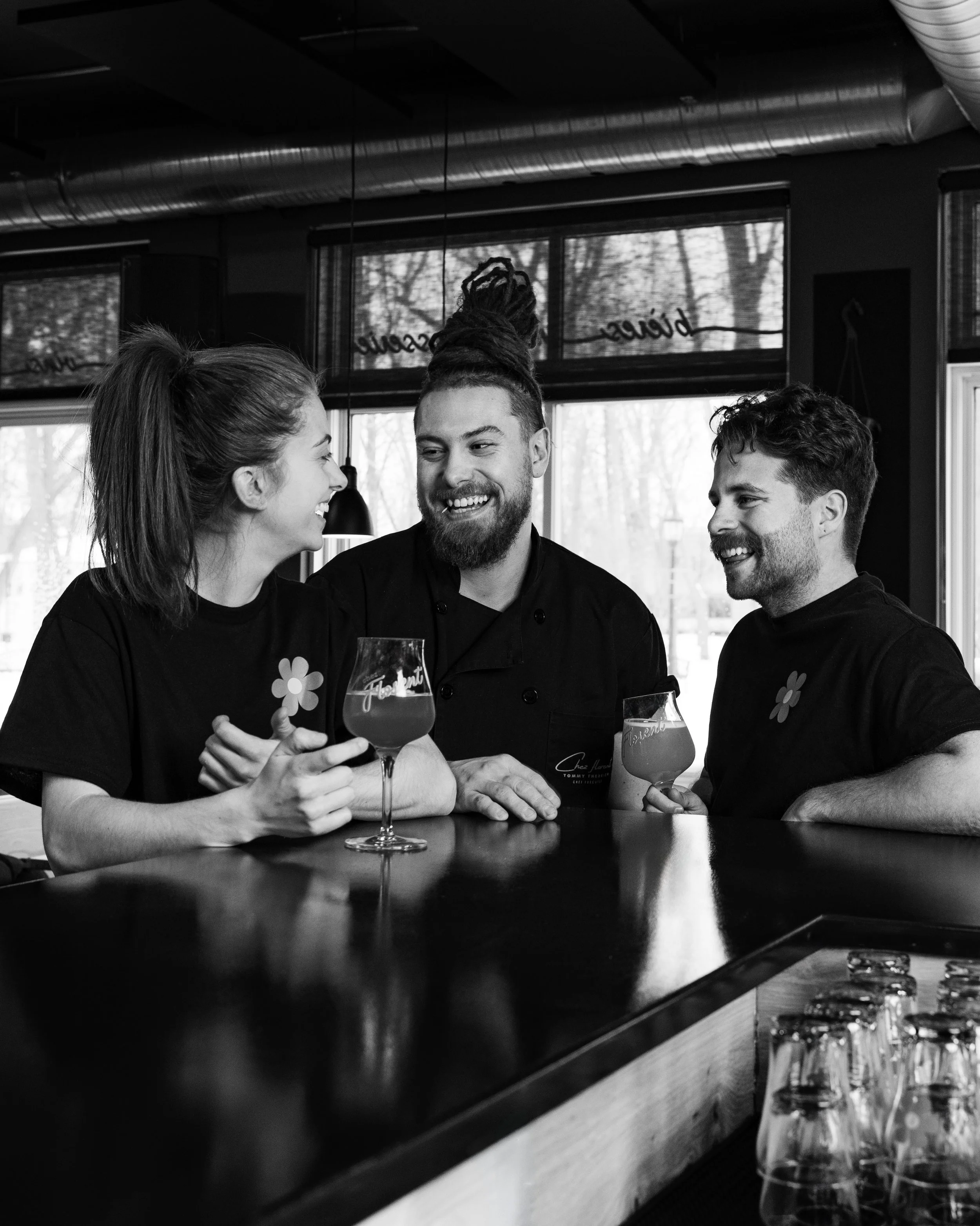 Three people smiling and laughing together at a bar or cafe, holding glasses of beer or a similar drink, with a window showing trees outside in the background.