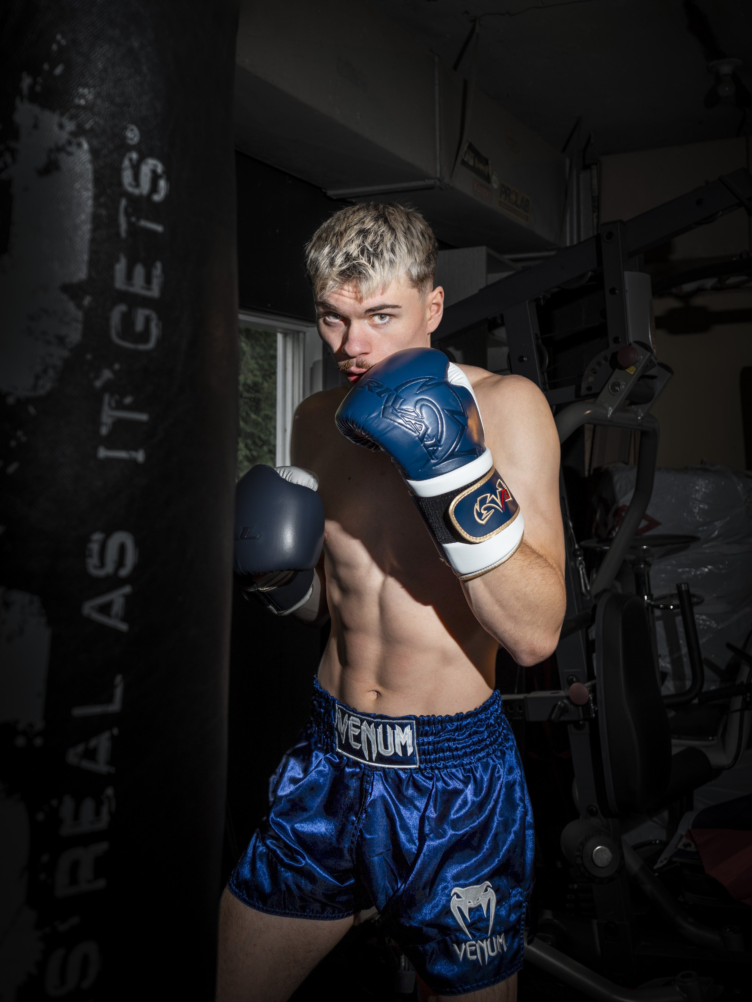 A young male boxer with short blond hair, wearing blue boxing shorts and gloves, practicing his punches in a gym. He is in a fighting stance near a black punching bag with white text.