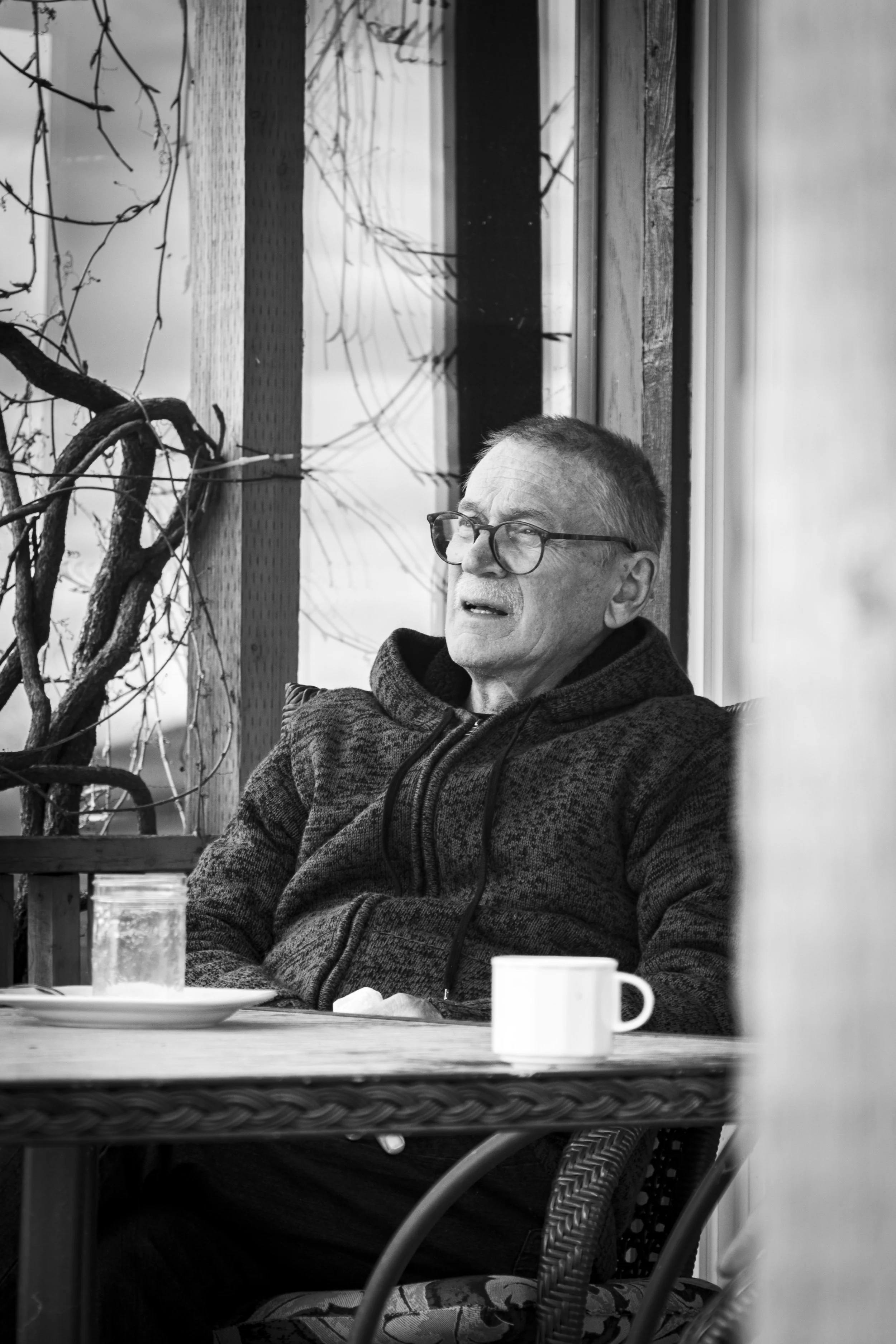 An elderly man with glasses, wearing a hoodie, sitting at a table with a plate and a mug, with a wooden wall and a leafless vine in the background.