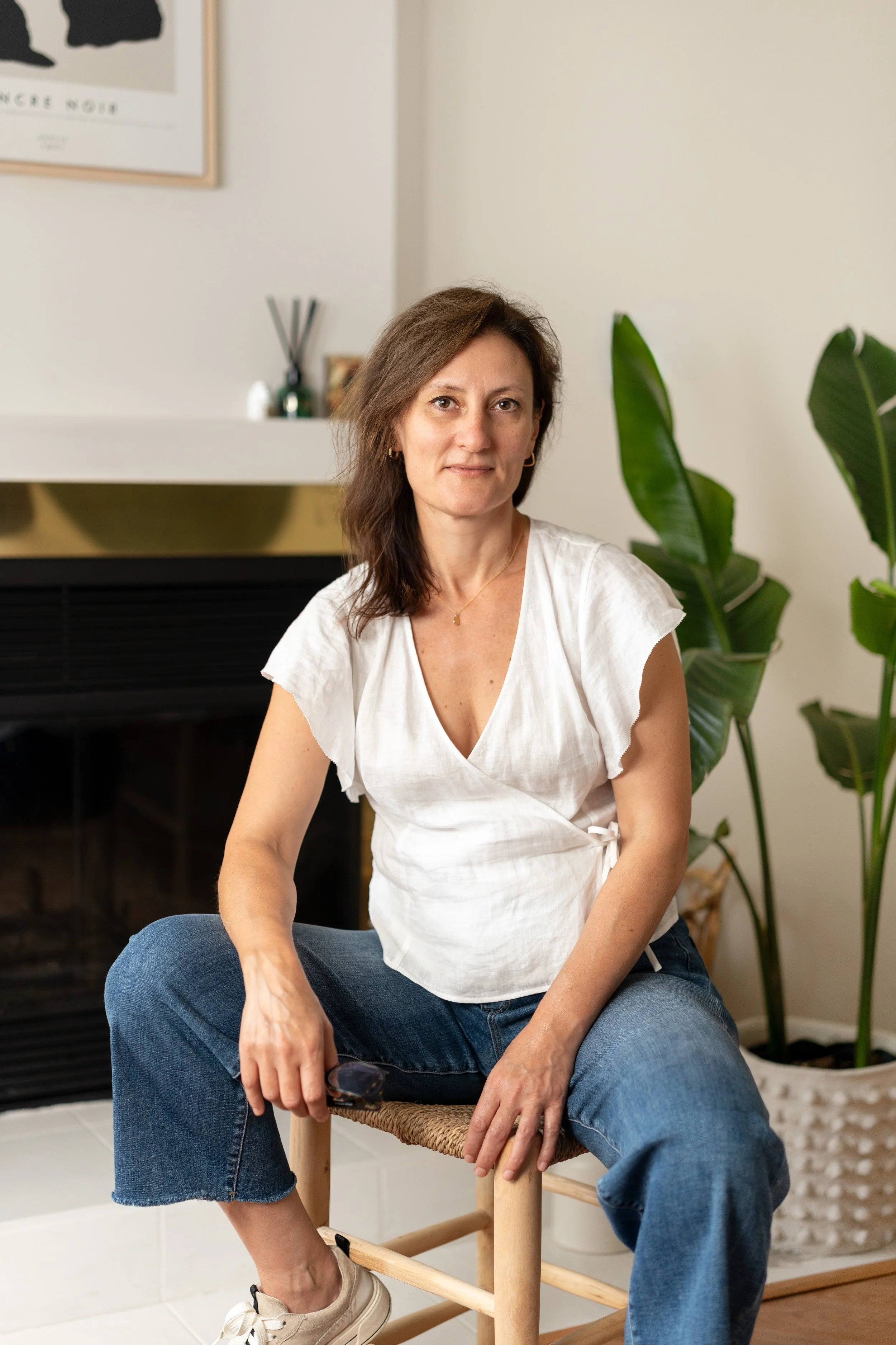 A woman with shoulder-length brown hair sitting on a woven chair in a living room. She is wearing a white top, blue jeans, and beige sneakers. There are green plants beside her and a fireplace behind her.