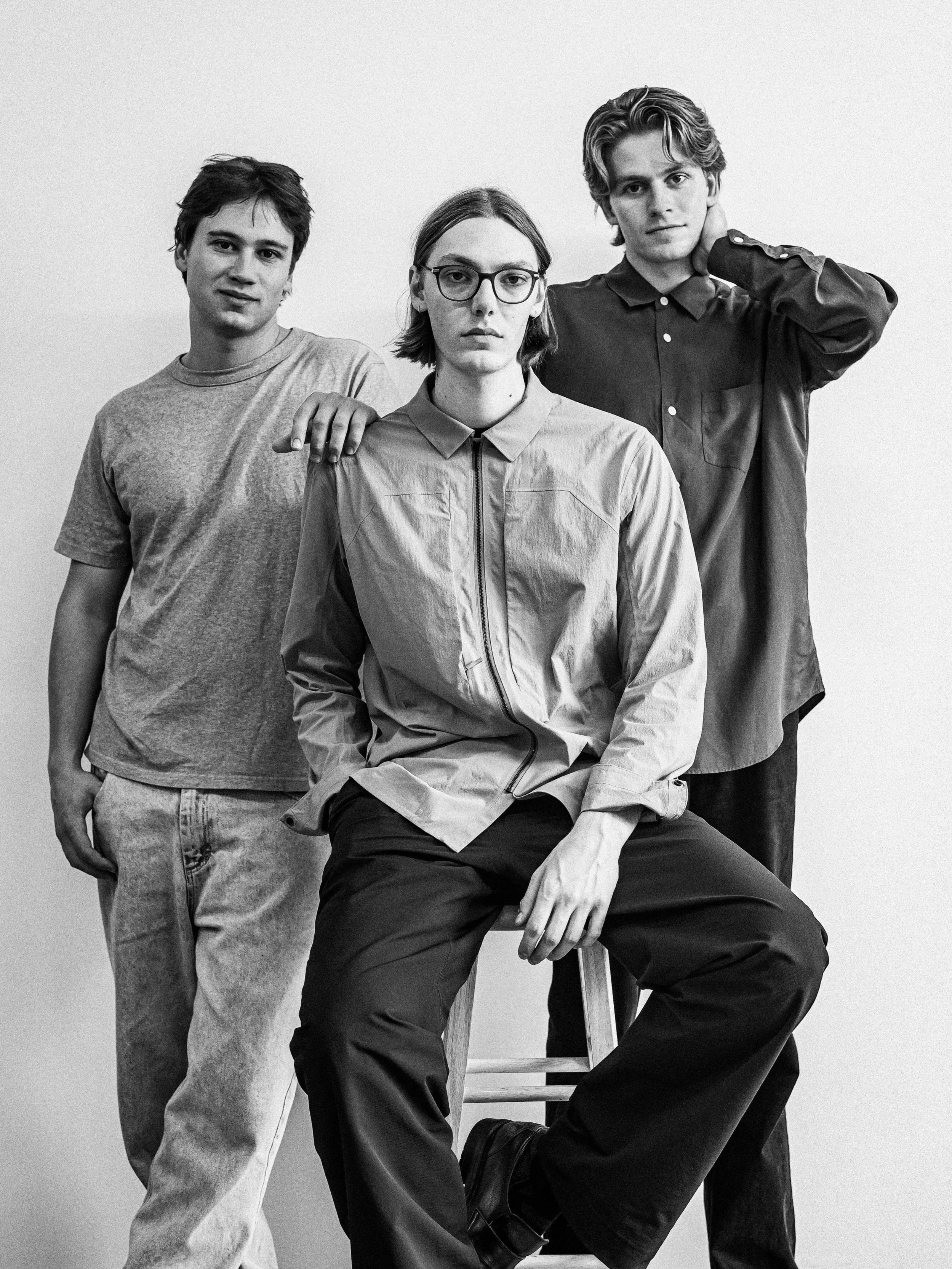 Black and white photo of three young adults, two men and one woman, posing together against a plain background. The woman is seated on a stool, wearing glasses and a light-colored jacket. The man on the left is standing with his hand on her shoulder, wearing a casual t-shirt and jeans. The man on the right is standing with one hand on the back of his neck, wearing a button-up shirt and dark pants.