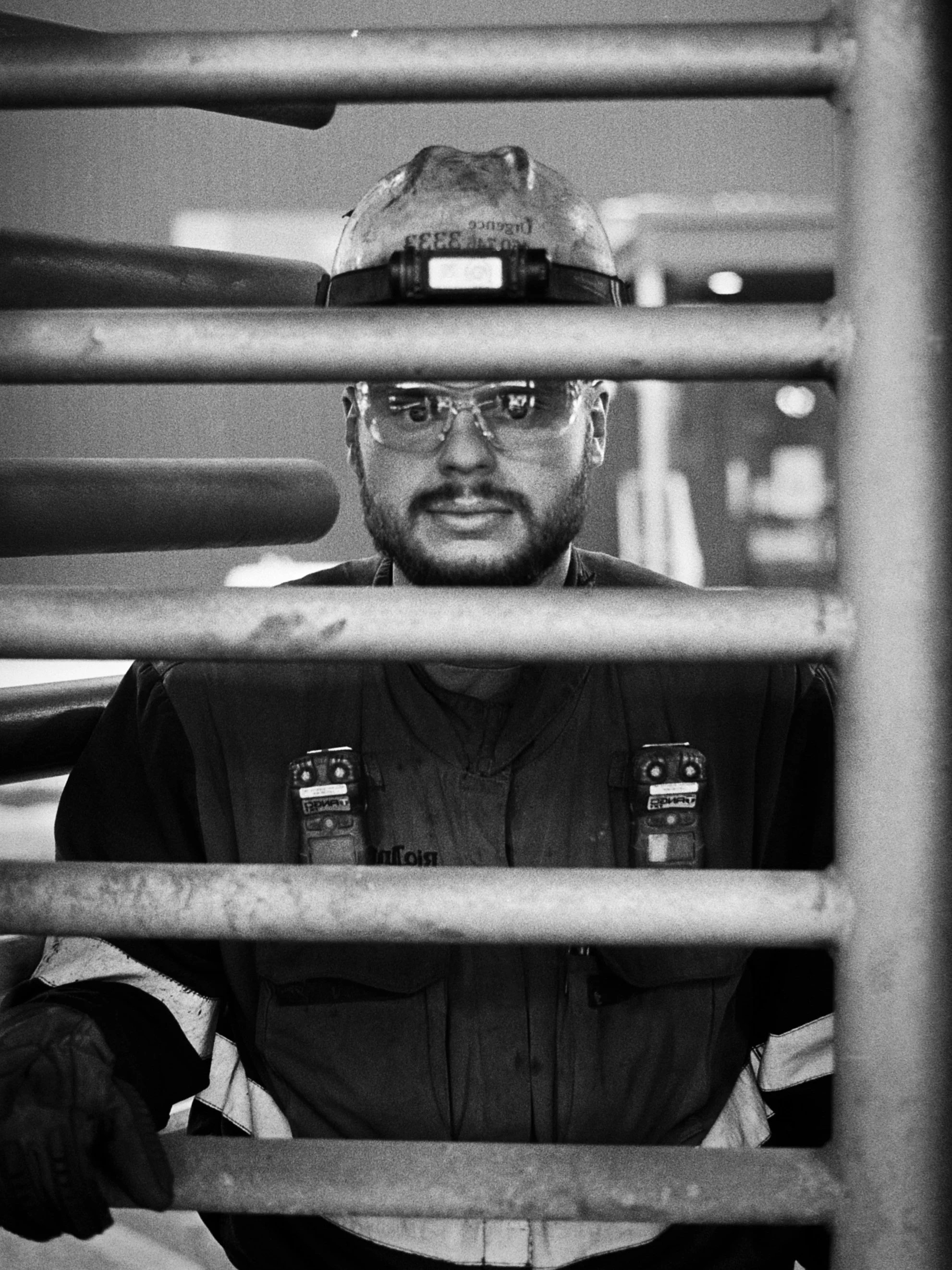 A black and white photo of a man taking a selfie through a mirror or a window with horizontal bars in front of him. He is wearing safety glasses, a cap, and a vest with various patches, suggesting he is at work in a technical or industrial setting.