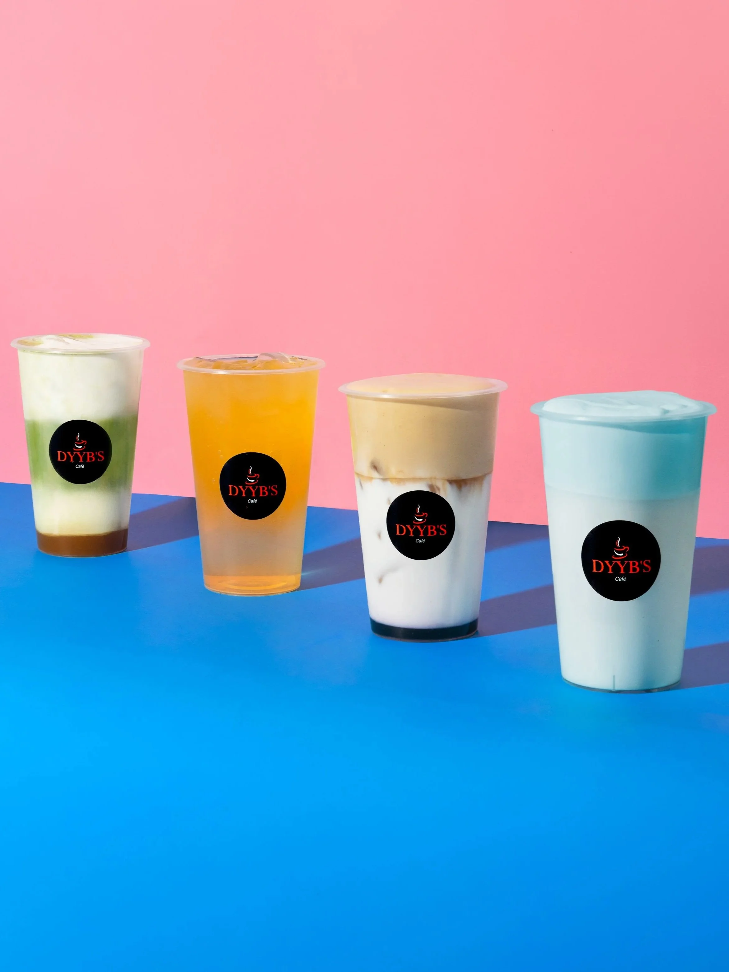 Four colorful cups with the logo 'DYYB'S Café' on a blue surface against a pink background.