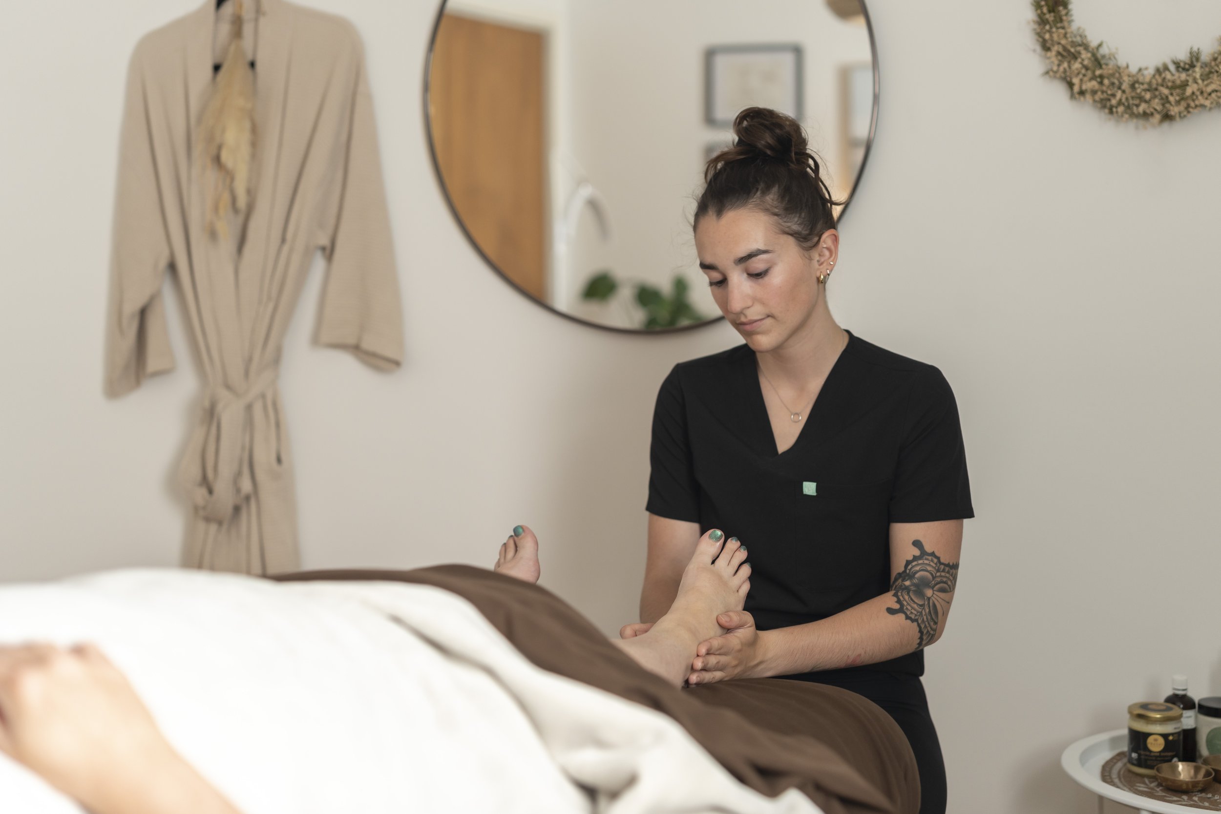 A woman with dark brown hair in a bun, wearing earrings, a black shirt, and displaying tattoos on her arm, is holding a person's foot in a room decorated with a mirror, a wicker wall hanging, and framed pictures.