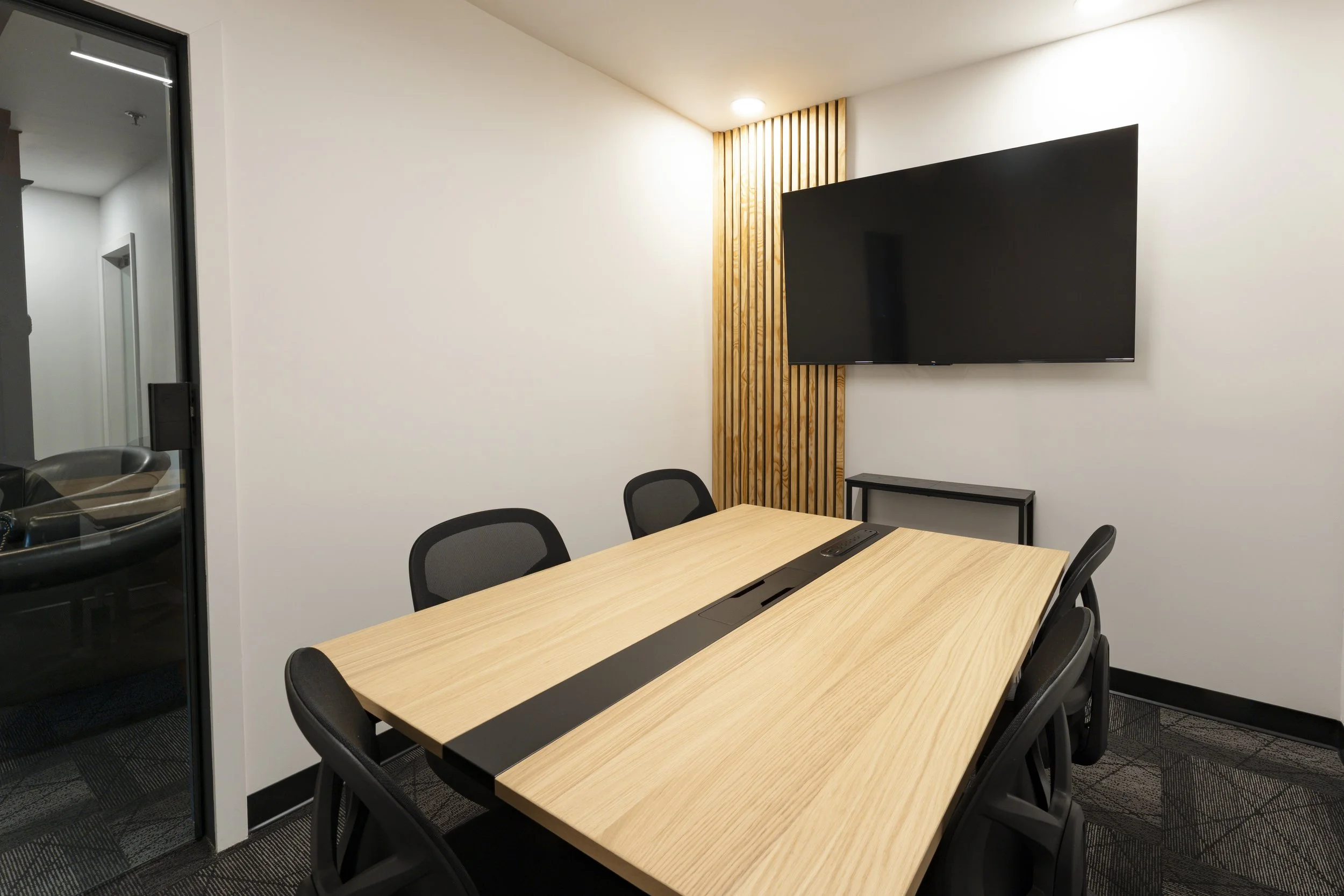 A modern conference room with a wooden table, black chairs, a wall-mounted flat-screen TV, and a decorative wood panel on the wall.