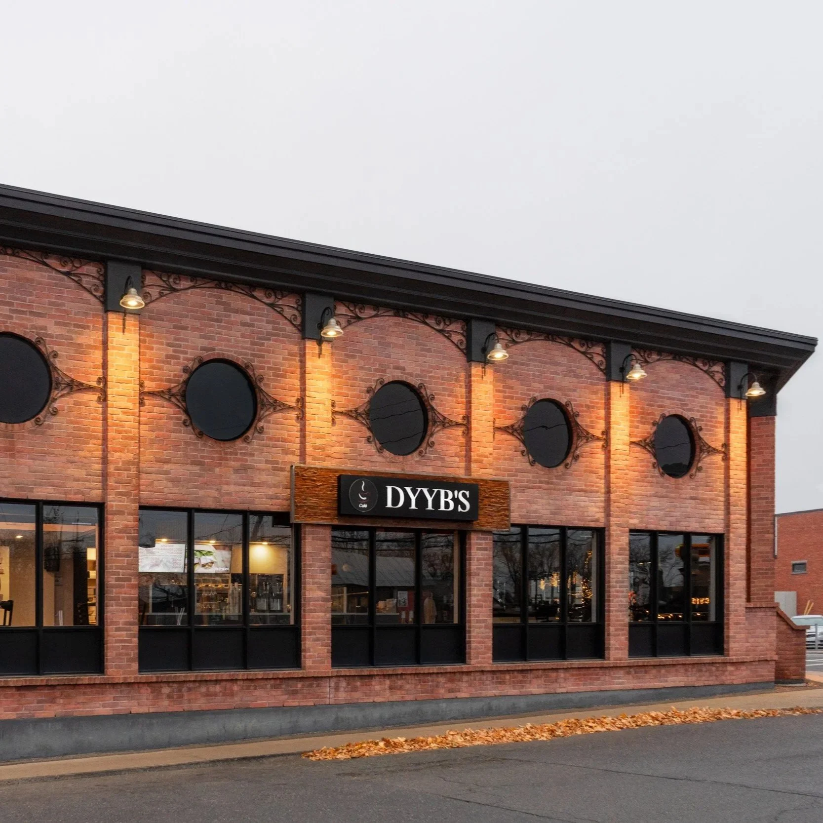 Front view of a two-story brick commercial building with large windows and a sign that reads 'DYBB'S'. The building has black decorative ironwork and mounted outdoor lights. The street in front has fallen leaves and a cloudy sky overhead.