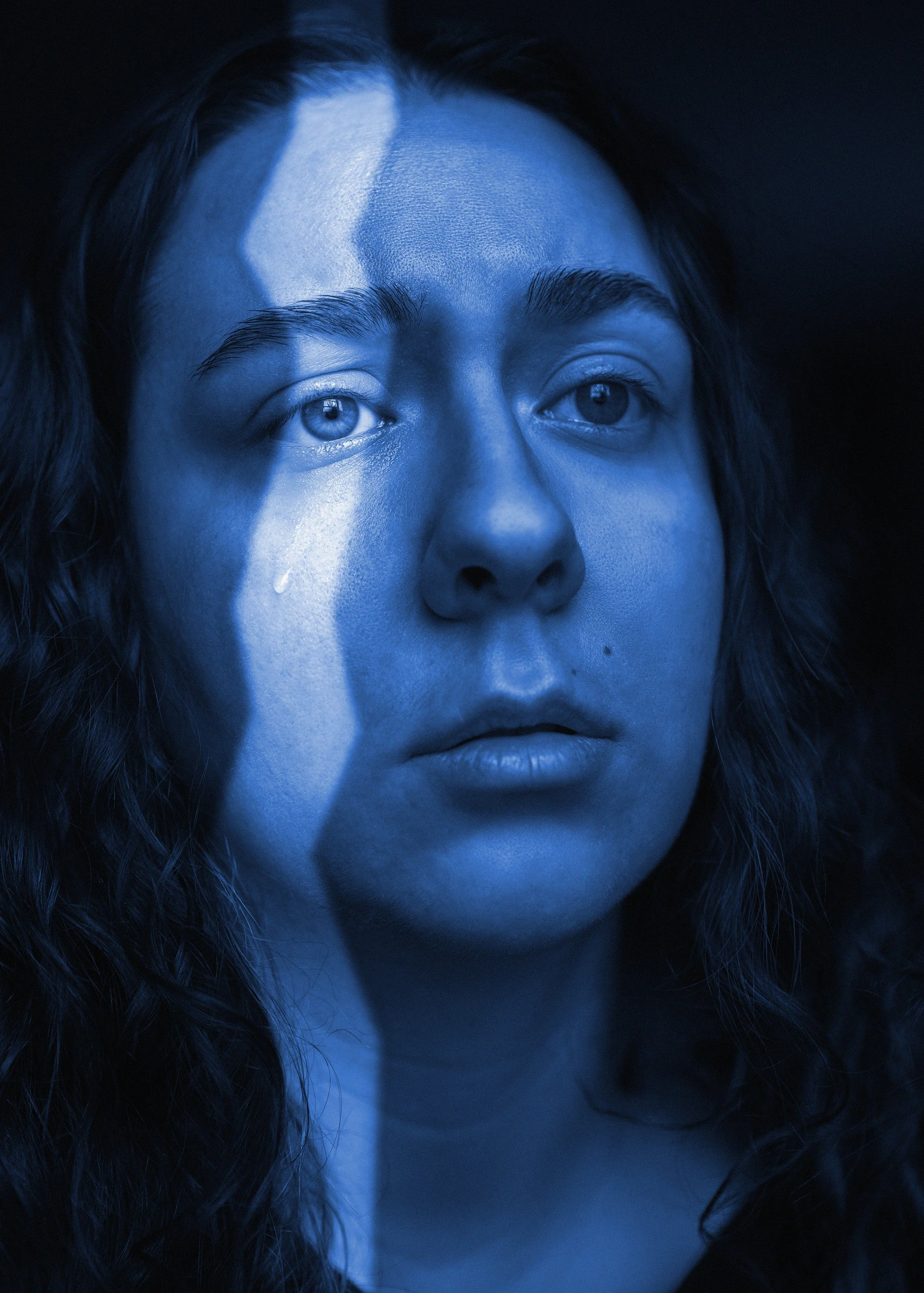 Close-up portrait of a woman's face illuminated with blue light, showing her eyes, nose, lips, and wavy hair against a dark background.