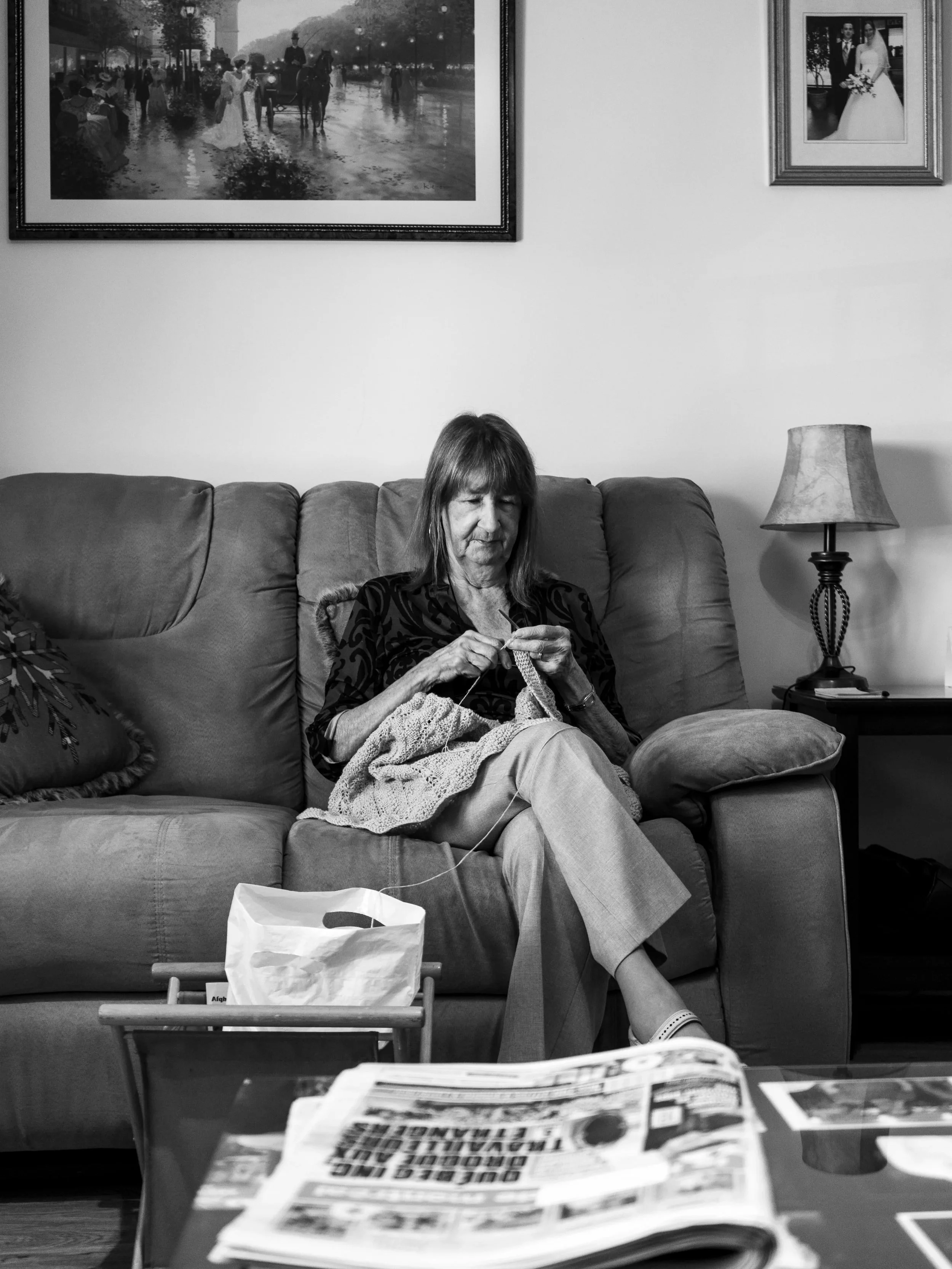A woman sitting on a couch knitting, with a table in front of her that has newspapers and a shopping bag, in a living room with a lamp and framed photographs on the wall.