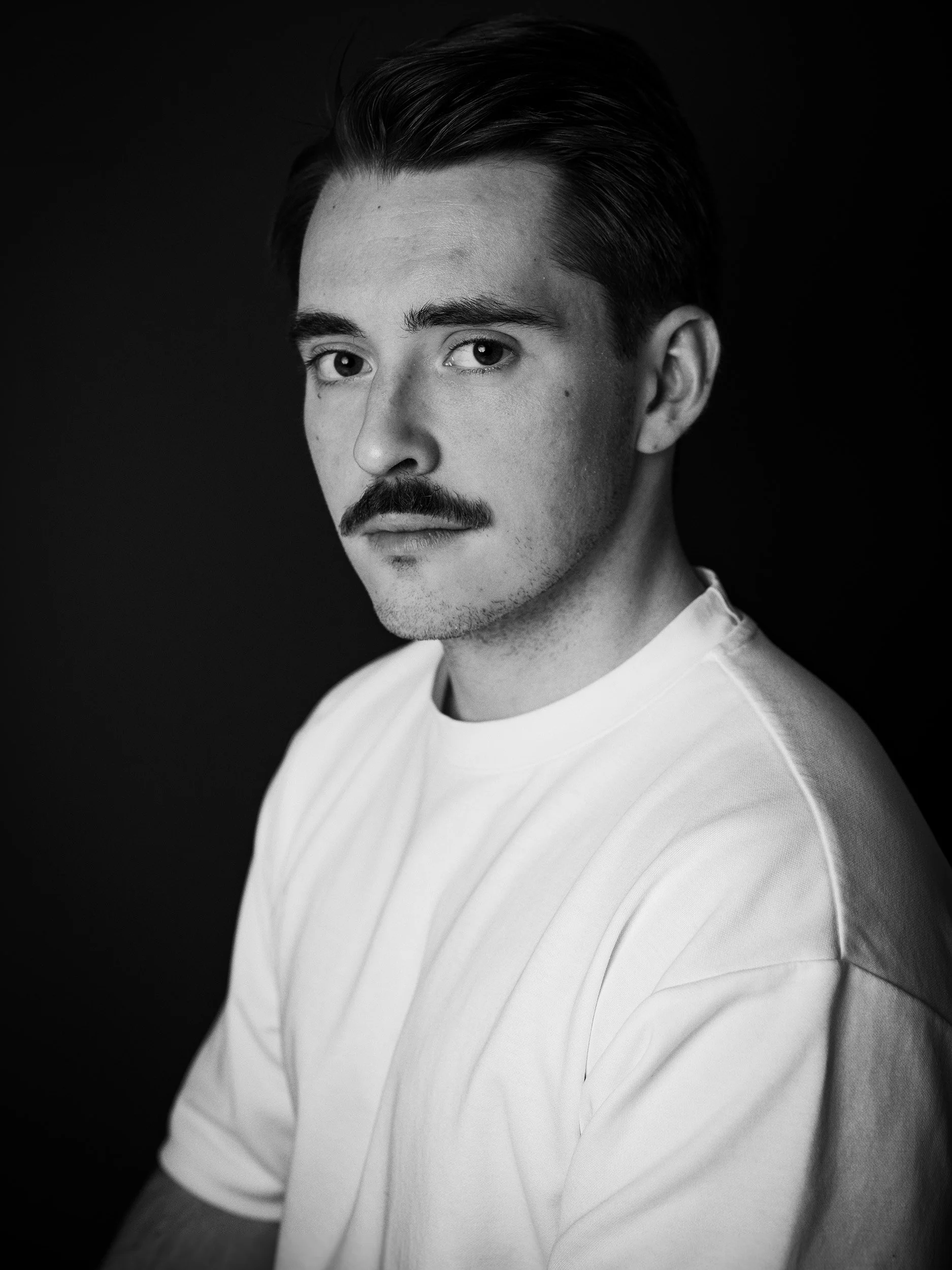 Black and white portrait of a young man with dark hair, mustache, and goatee, wearing a white shirt, looking directly at the camera against a dark background.