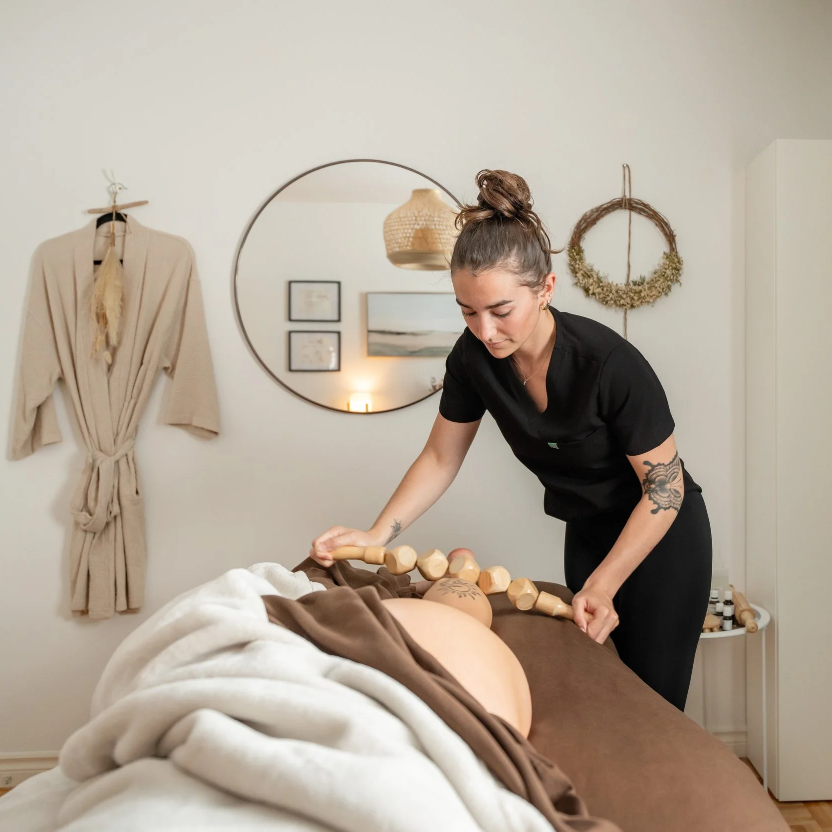 A woman in black uniform giving a massage on a client's back with wooden massage tools in a spa setting, decorated with wall art and a beige robe hanging on the wall.