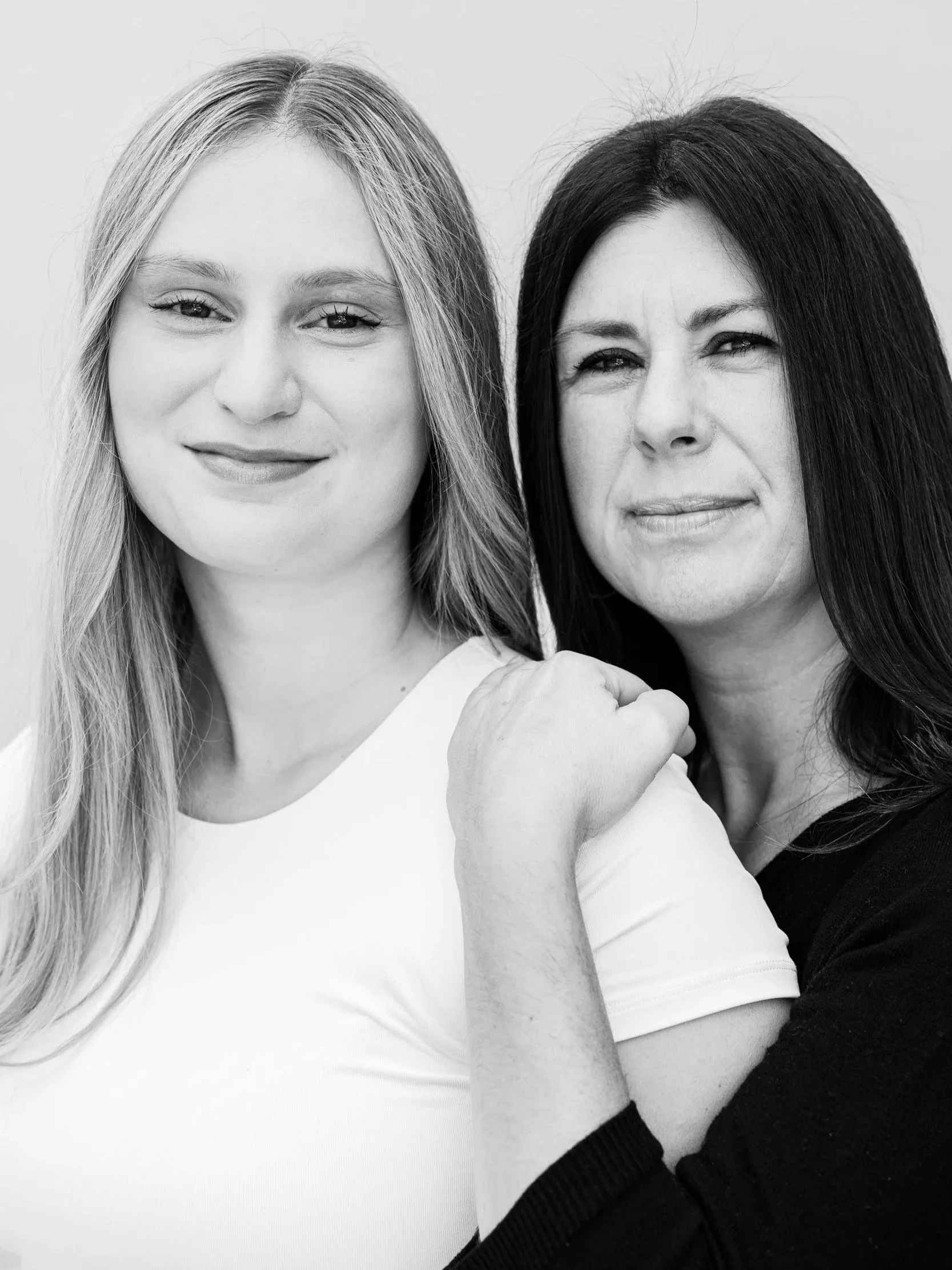 A black and white photo of two women, one with long blonde hair and the other with long dark hair, smiling at the camera, with the woman on the right having her arm around the one on the left.