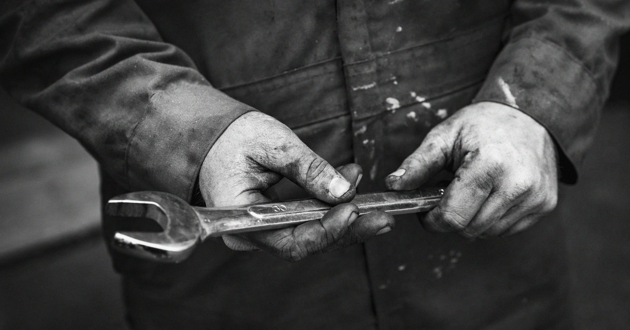 Close-up of a worker's hands holding a wrench, showing rough, dirty hands and worn clothing with paint splatters.