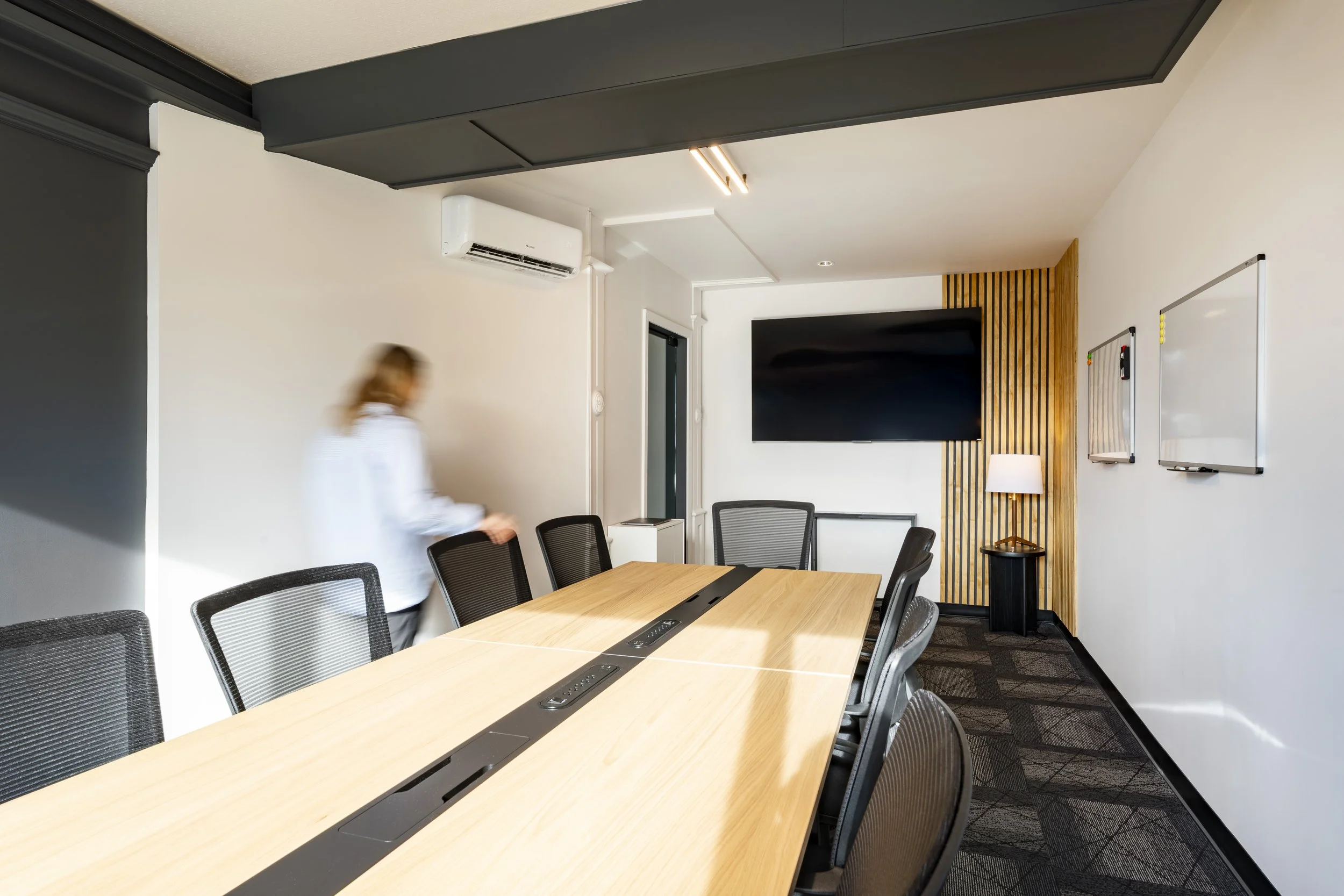 A modern conference room with a long wooden table, black mesh chairs, a flat-screen TV on a white wall, and two whiteboards, with a blurred person walking through the room.