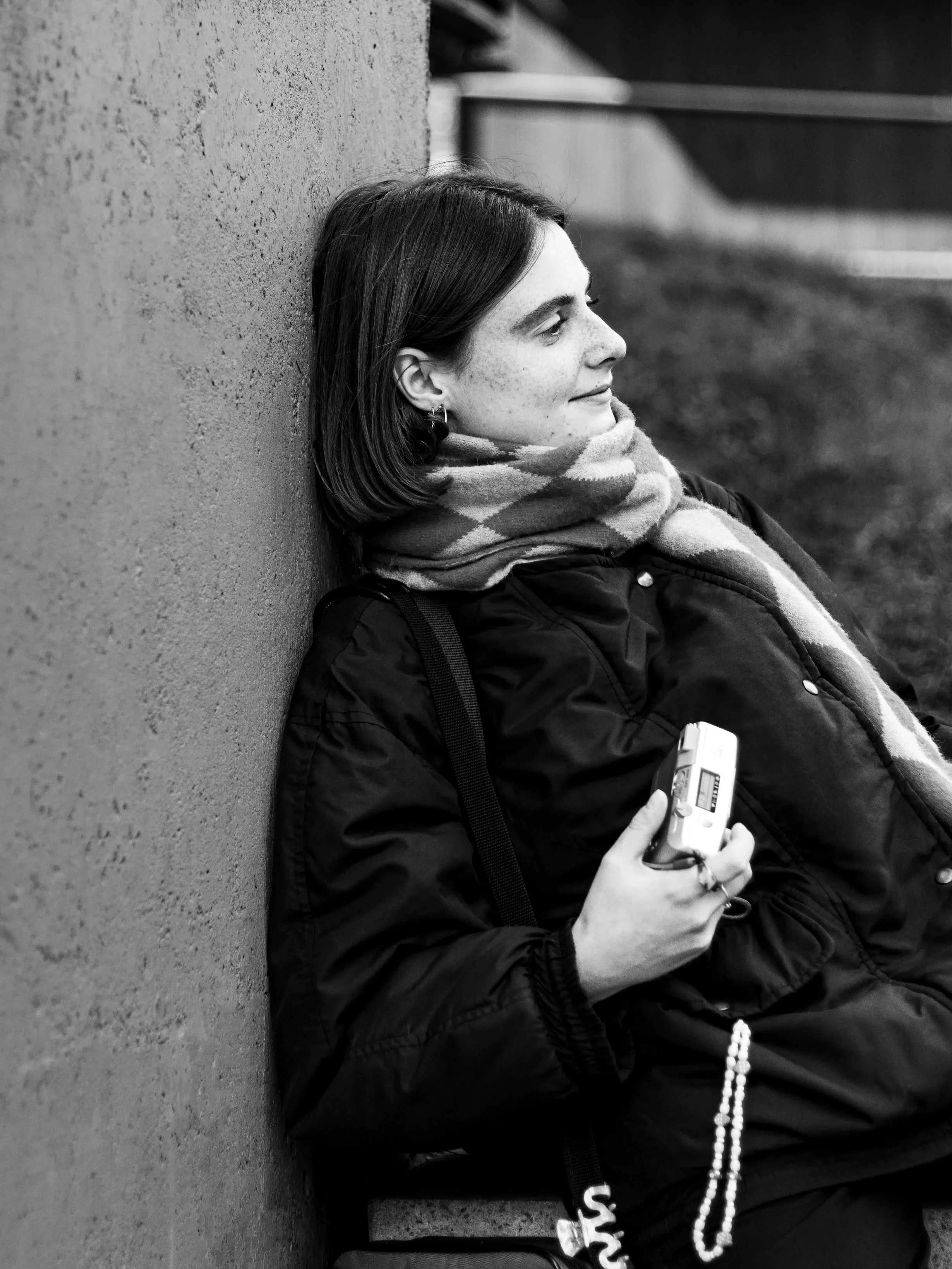 A woman with dark hair and freckles sitting with her back against a wall, looking to her right, holding a small electronic device in her hand, wearing a scarf and a dark jacket.