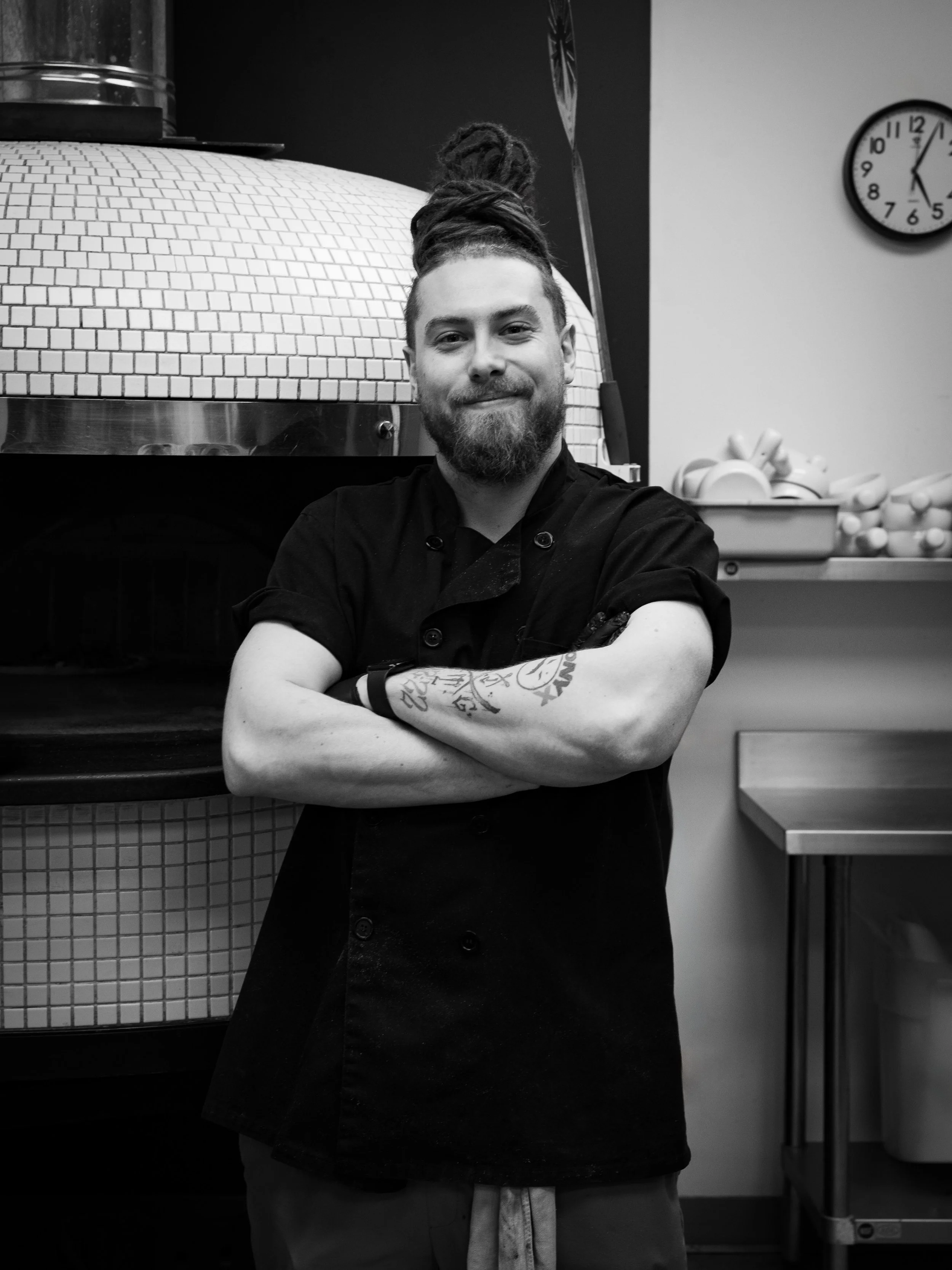 A man with a beard and dreadlocks standing with arms crossed in a kitchen or restaurant setting, wearing a black chef's coat.