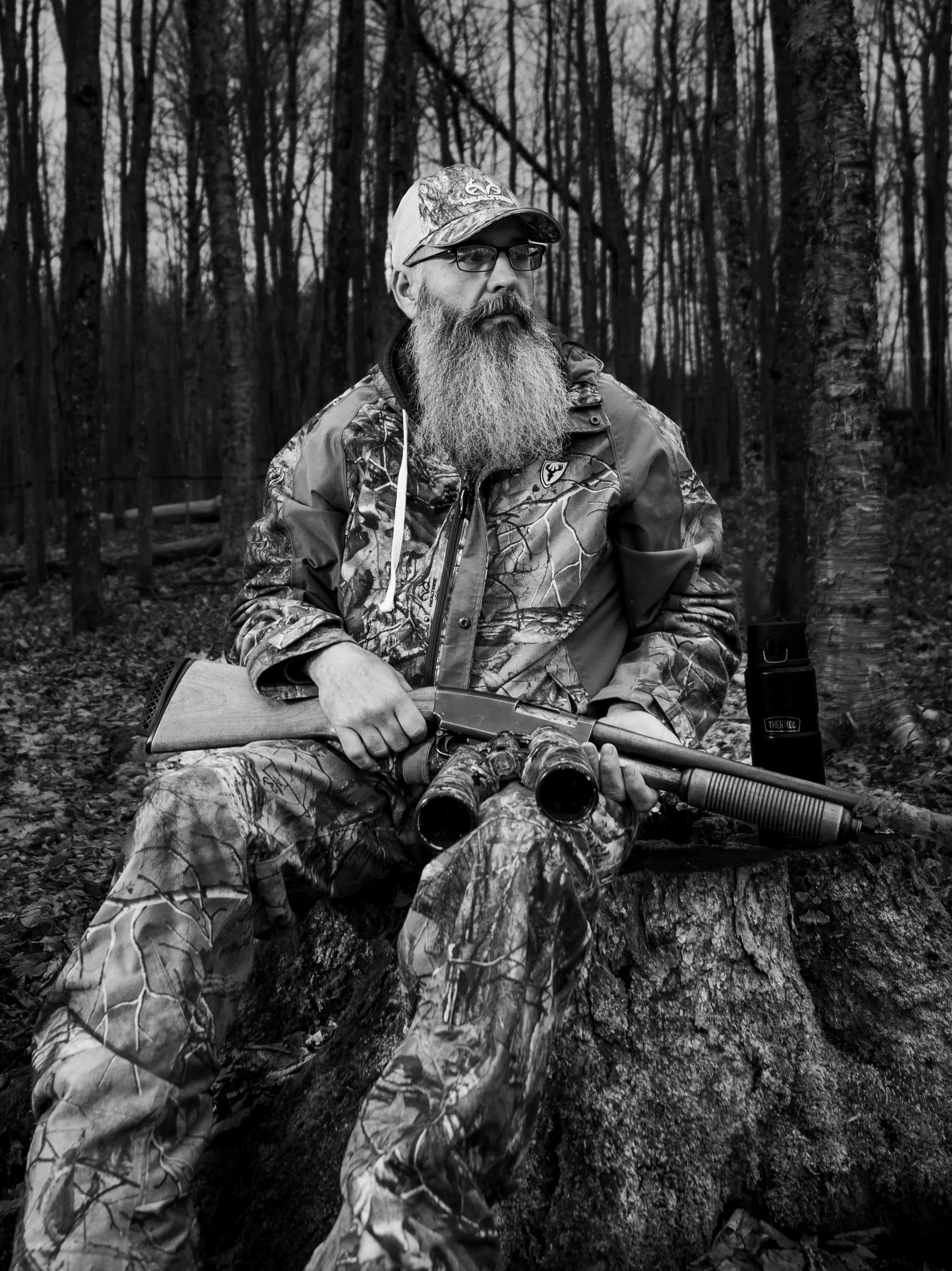 A man with a long beard and glasses sitting on a fallen log in a forest, holding a rifle with a scope, surrounded by leafless trees in black and white.