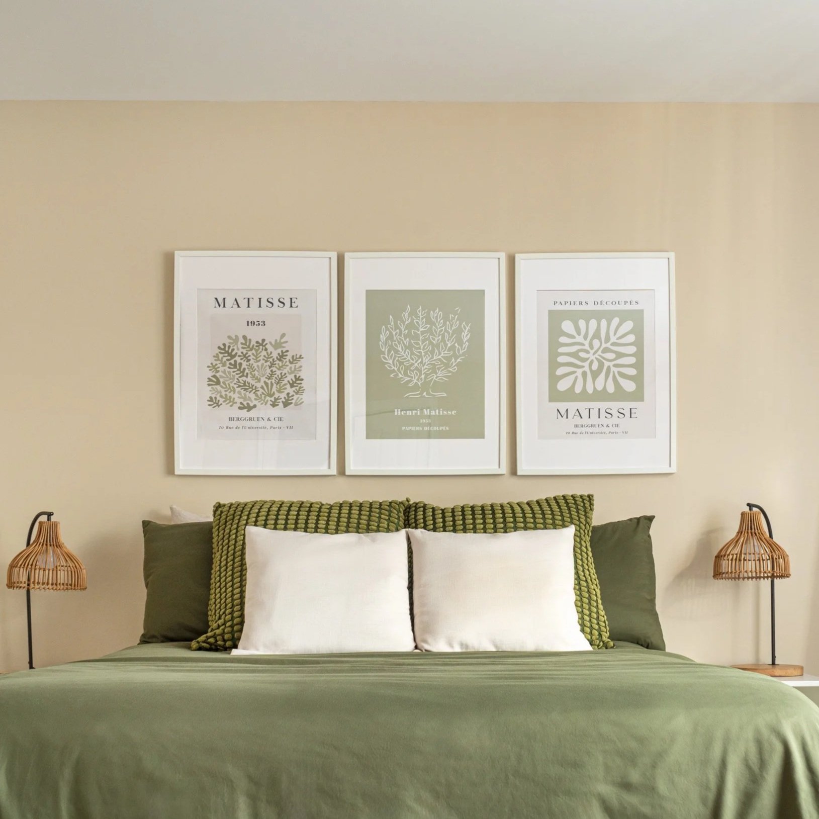 A bedroom with a bed covered in a green bedspread, topped with white and green pillows. Above the bed are three framed botanical prints on a beige wall, and on each side of the bed are rattan night lamps.