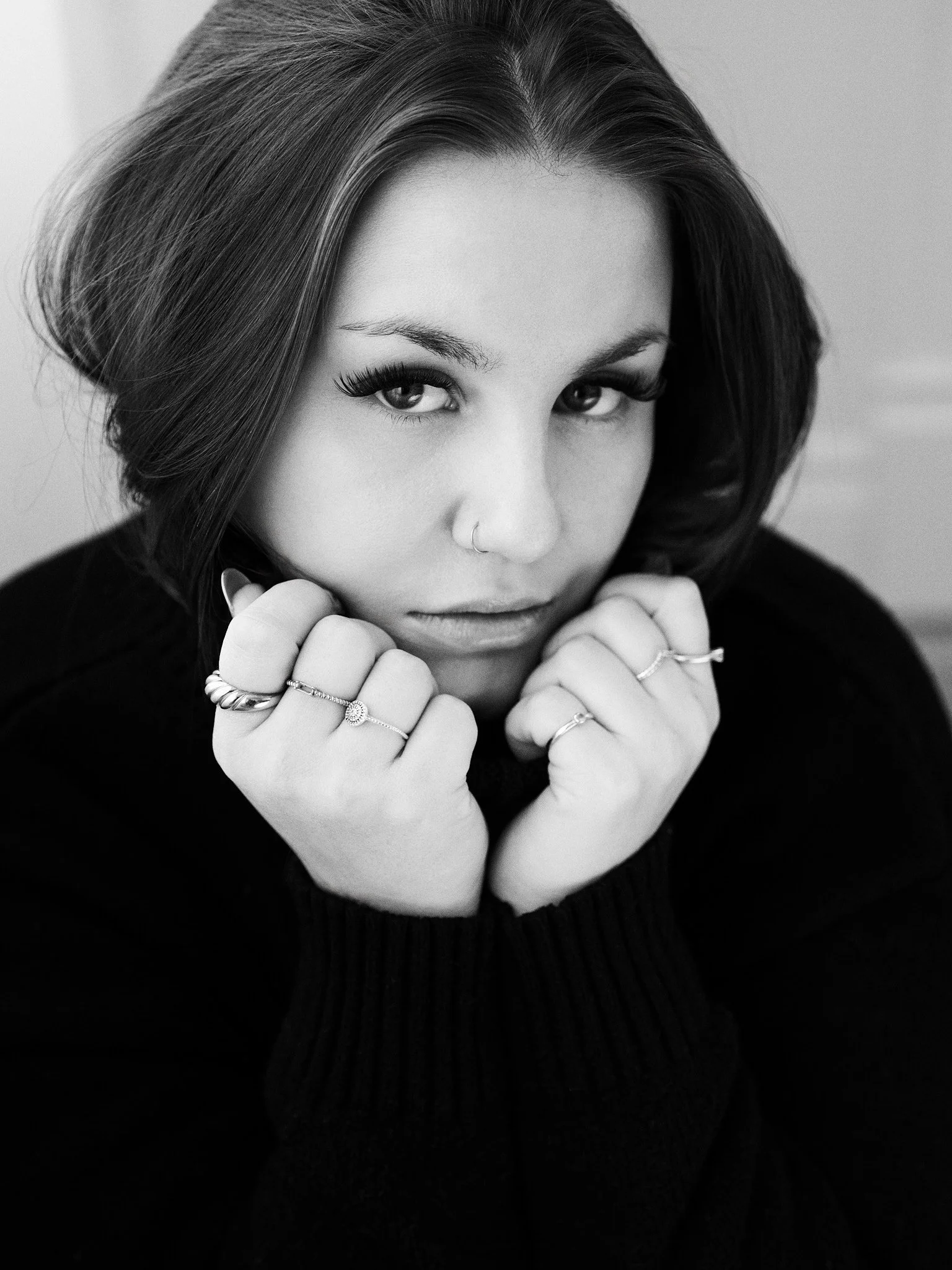 Black and white portrait of a woman with dark hair, wearing multiple rings, looking directly at the camera with her hands near her chin.