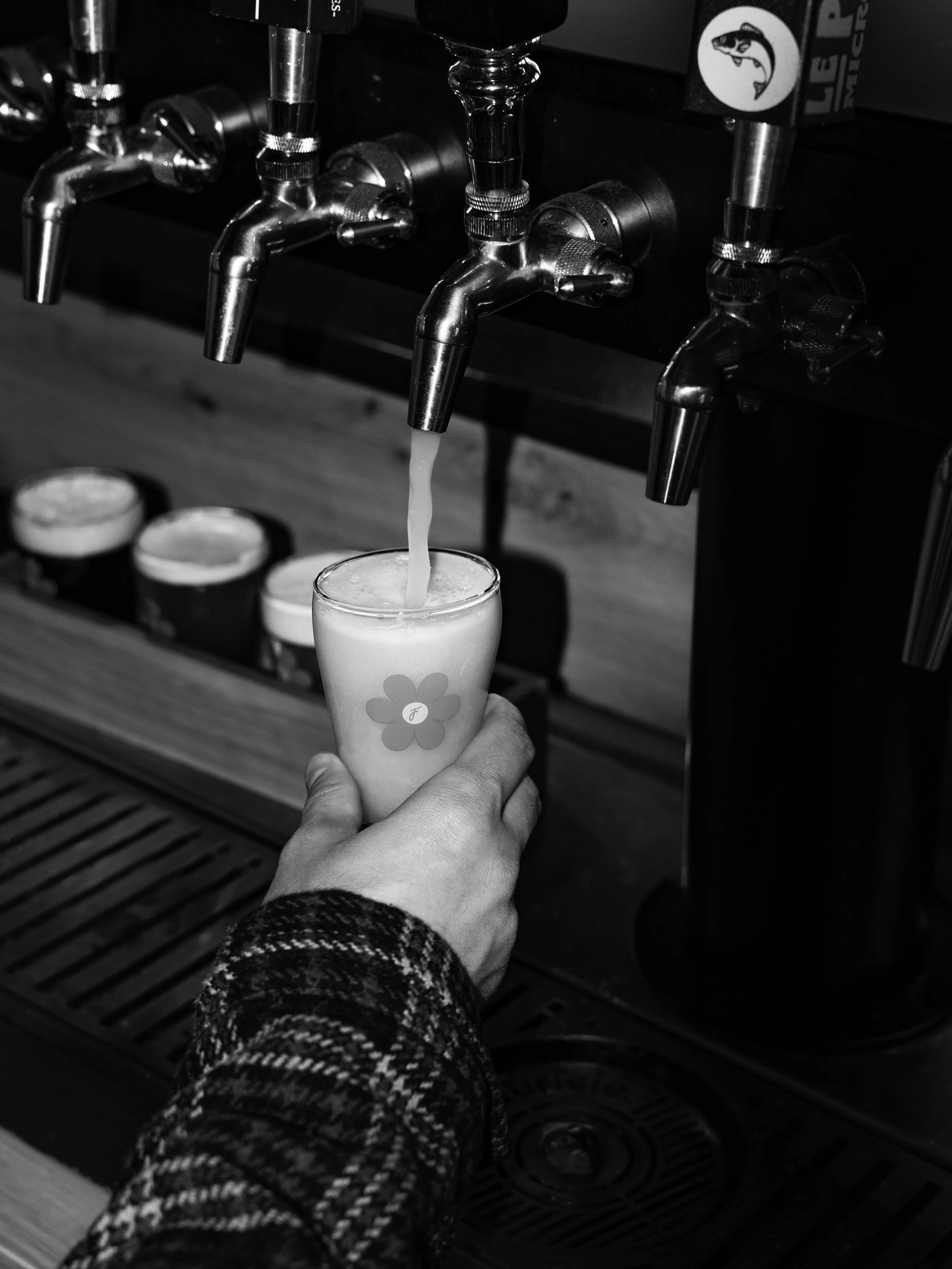 A person fills a glass with a beverage from a chrome tap at a bar or brewery, with a logo of a fish on the tap.