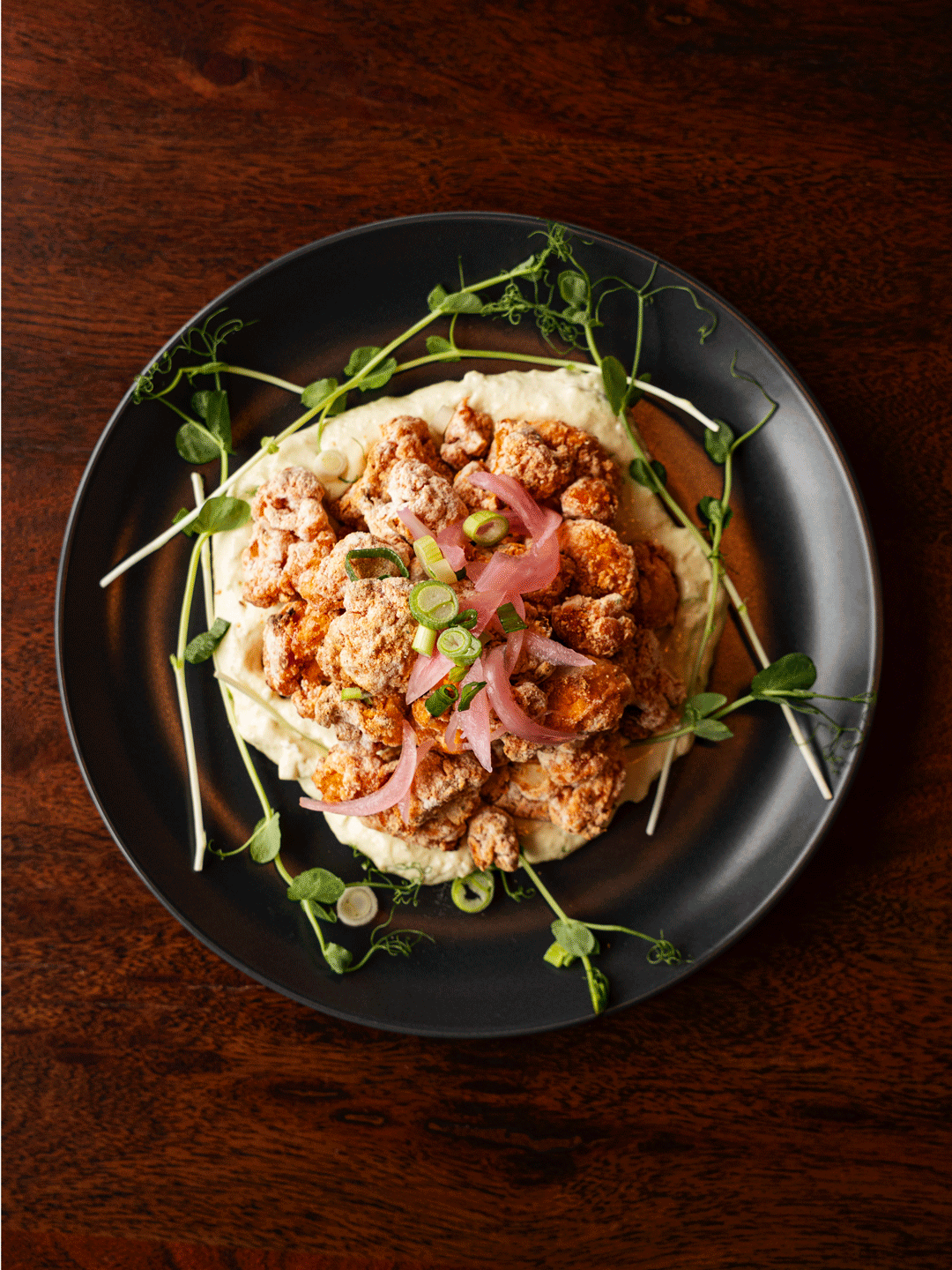 Plate of fried chicken pieces topped with sliced green onions and pickled red onions, served over mashed potatoes with green sprigs around the edge, on a dark plate on a wooden table.
