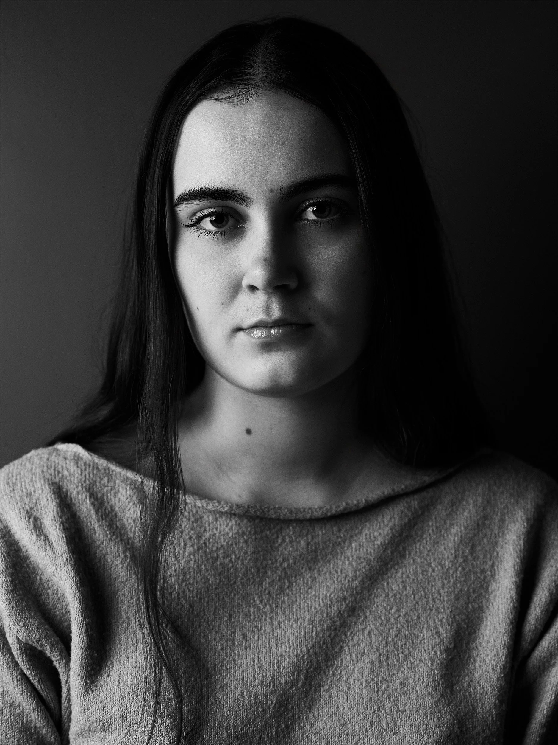 Black and white close-up portrait of a young woman with long dark hair and light-colored eyes, wearing a cozy sweater.