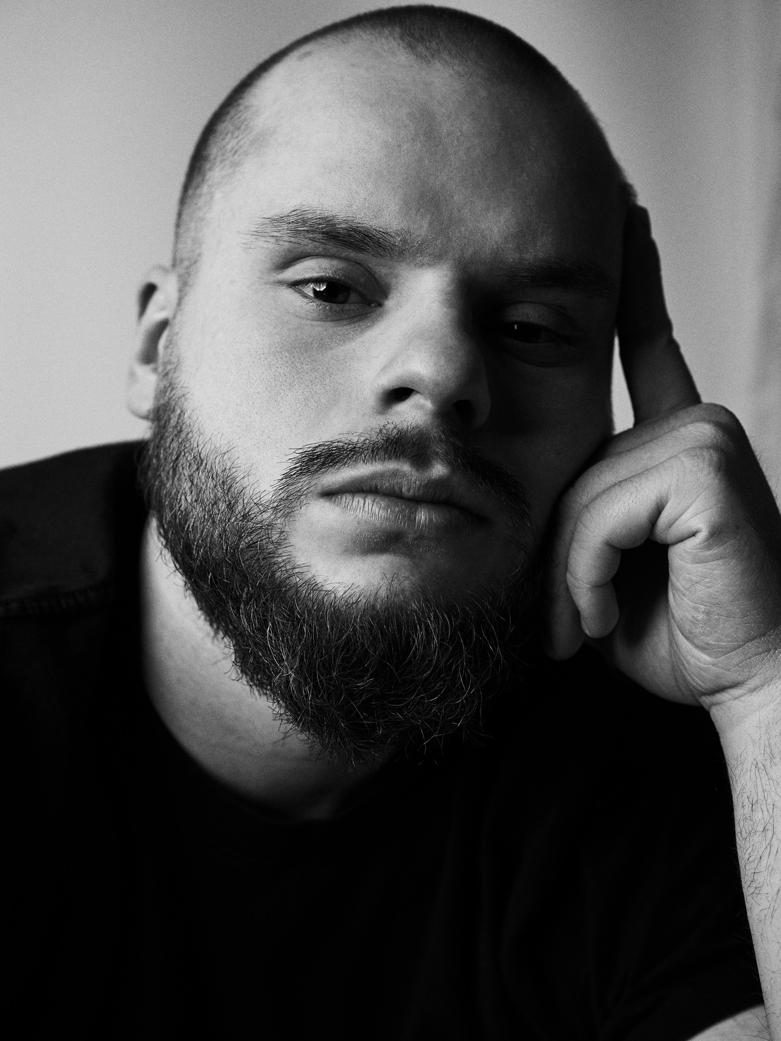 Black and white close-up photograph of a person with a shaved head and beard, resting their head on their hand with a contemplative expression.