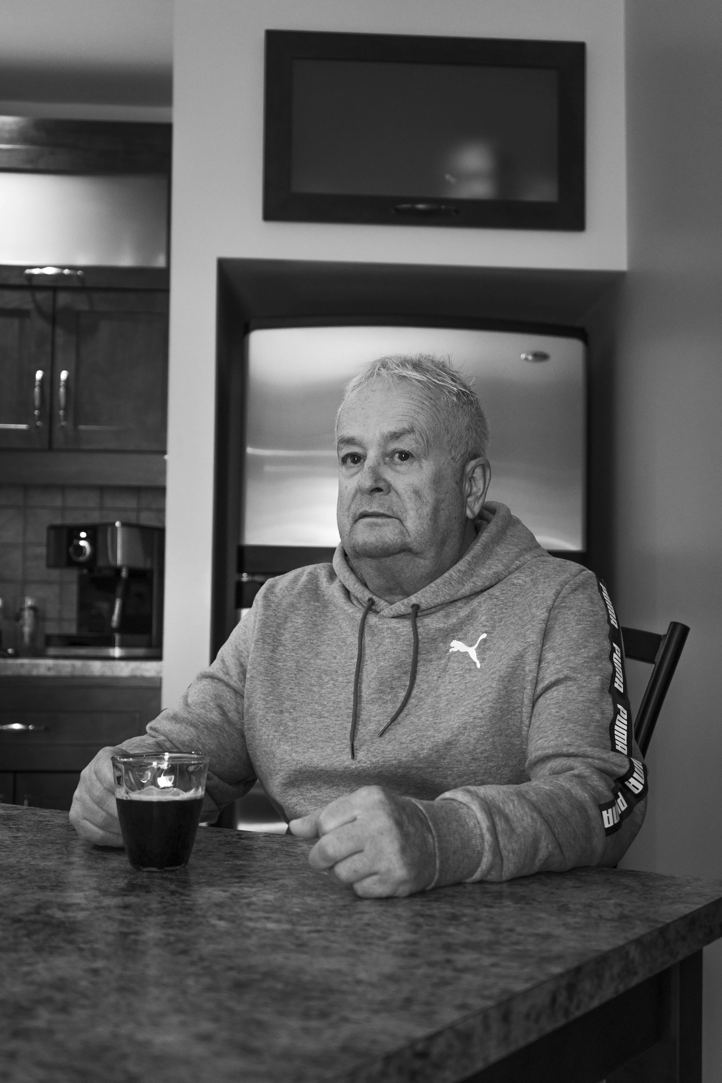 A middle-aged man with gray hair sitting at a table in a kitchen, holding a glass of dark beverage, wearing a gray hoodie, with a serious expression.