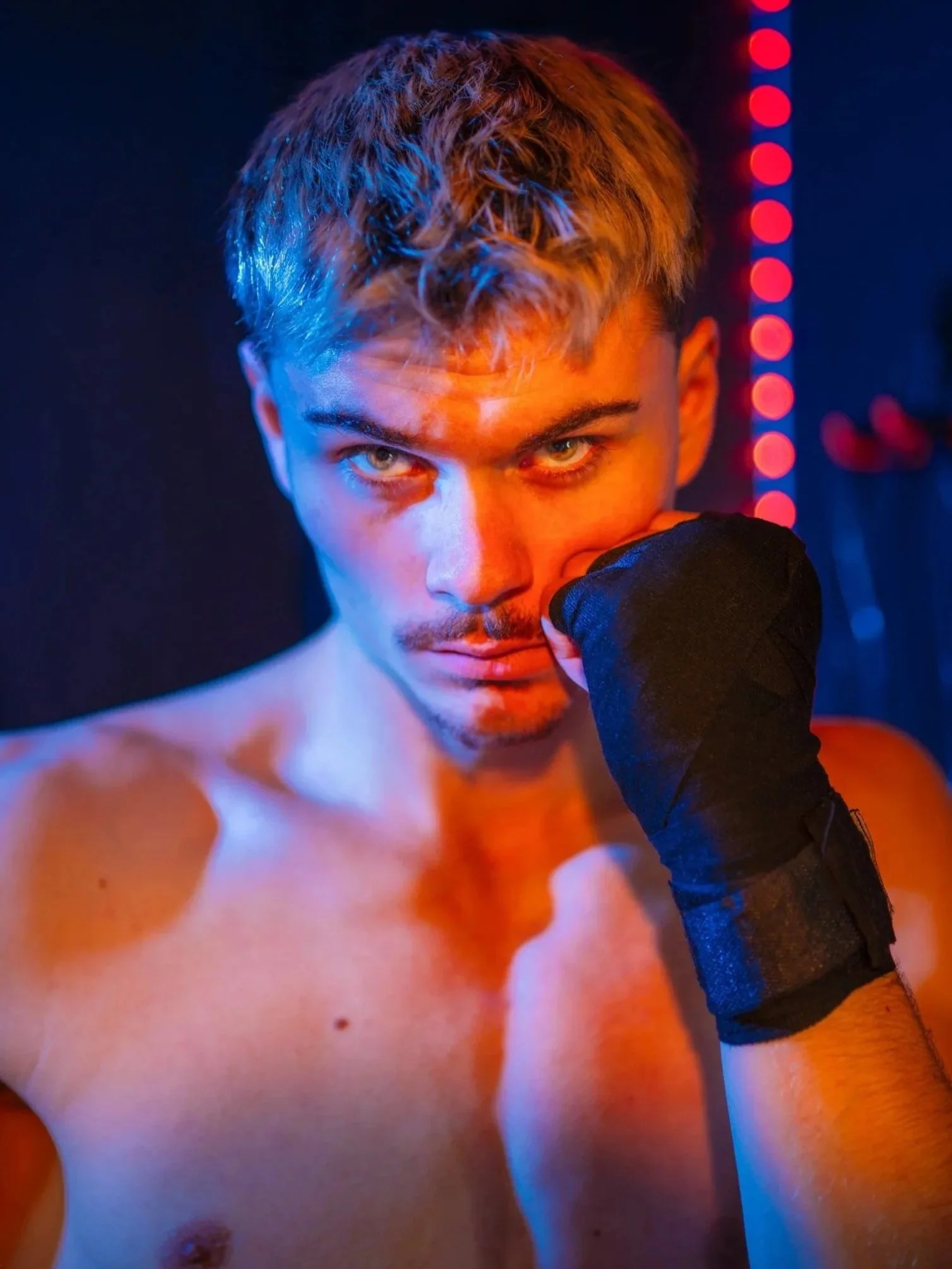Close-up of a young man with short hair and intense blue eyes, partially covering his face with a gloved hand, against a dark background with red and blue neon lights.