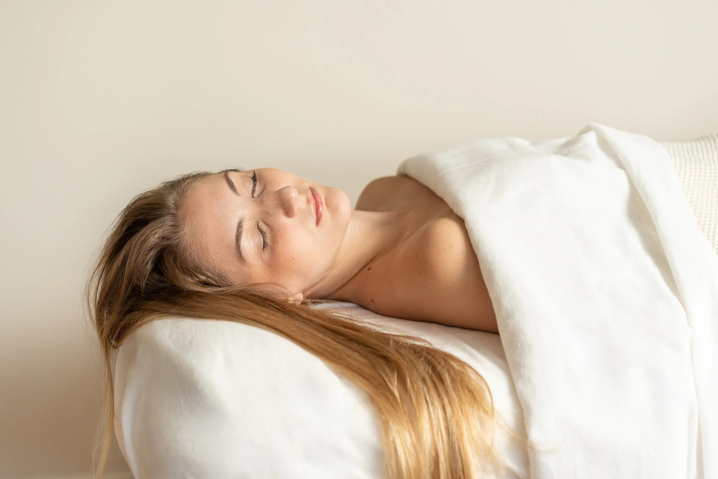 A woman lying on a bed with her eyes closed, covered with a white blanket, in a peaceful and relaxed state.