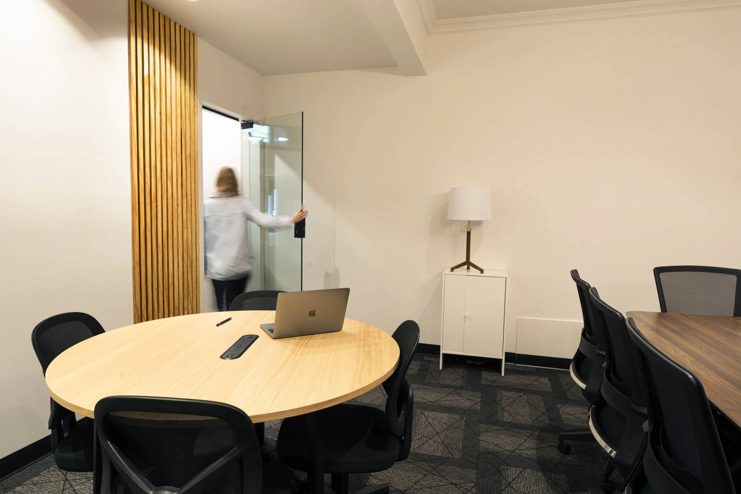 A modern conference room with a large wooden table and black office chairs. The room has white and dark gray walls, a wall-mounted flat-screen TV, and arched windows reflecting city lights at night.