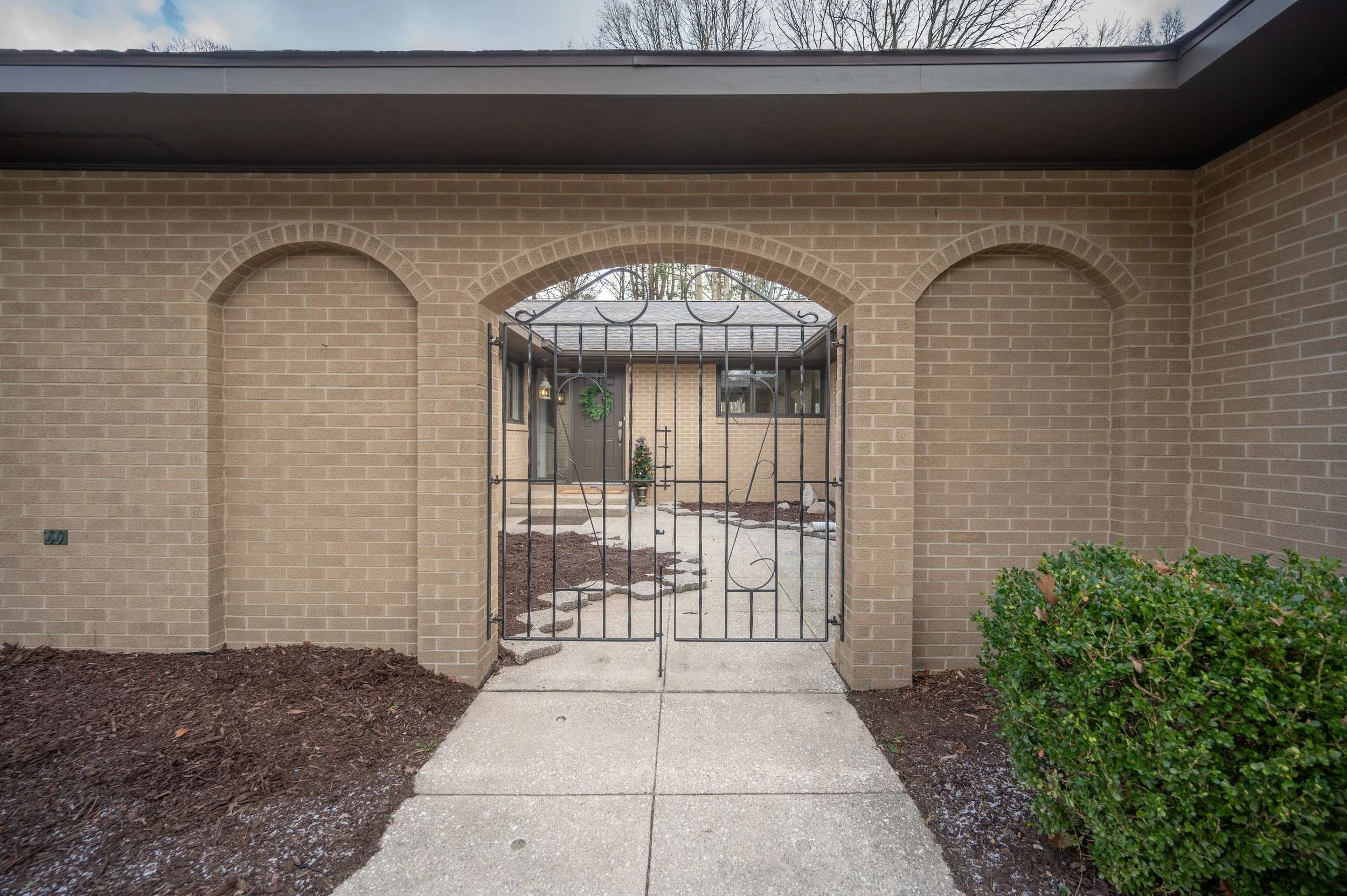 View of a brick wall and archway with a black gate leading to a house entrance, decorated with a green wreath, with shrubs on the sides and a concrete sidewalk in front.