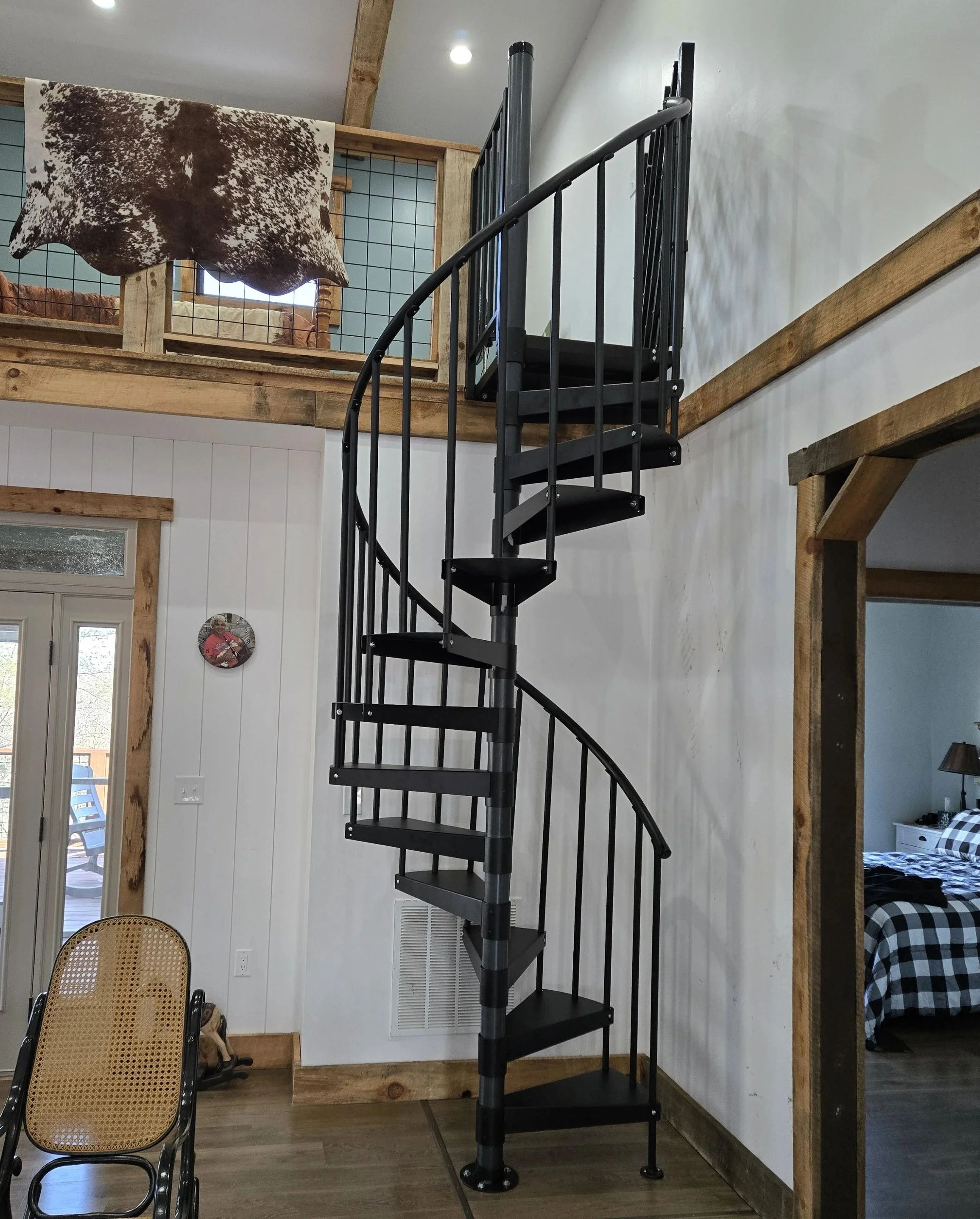 Interior of a house with a black spiral staircase, wooden trim, a chair with a cane back, and a loft area with a cowhide blanket.